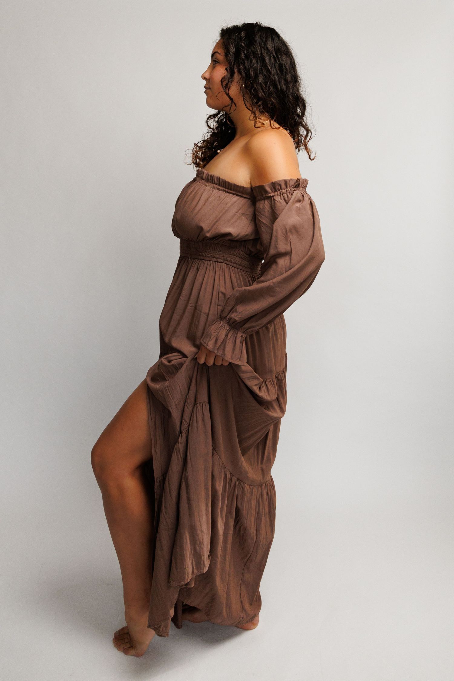 Woman in brown off-shoulder maxi dress with long sleeves and side slit, posing sideways on gray background.