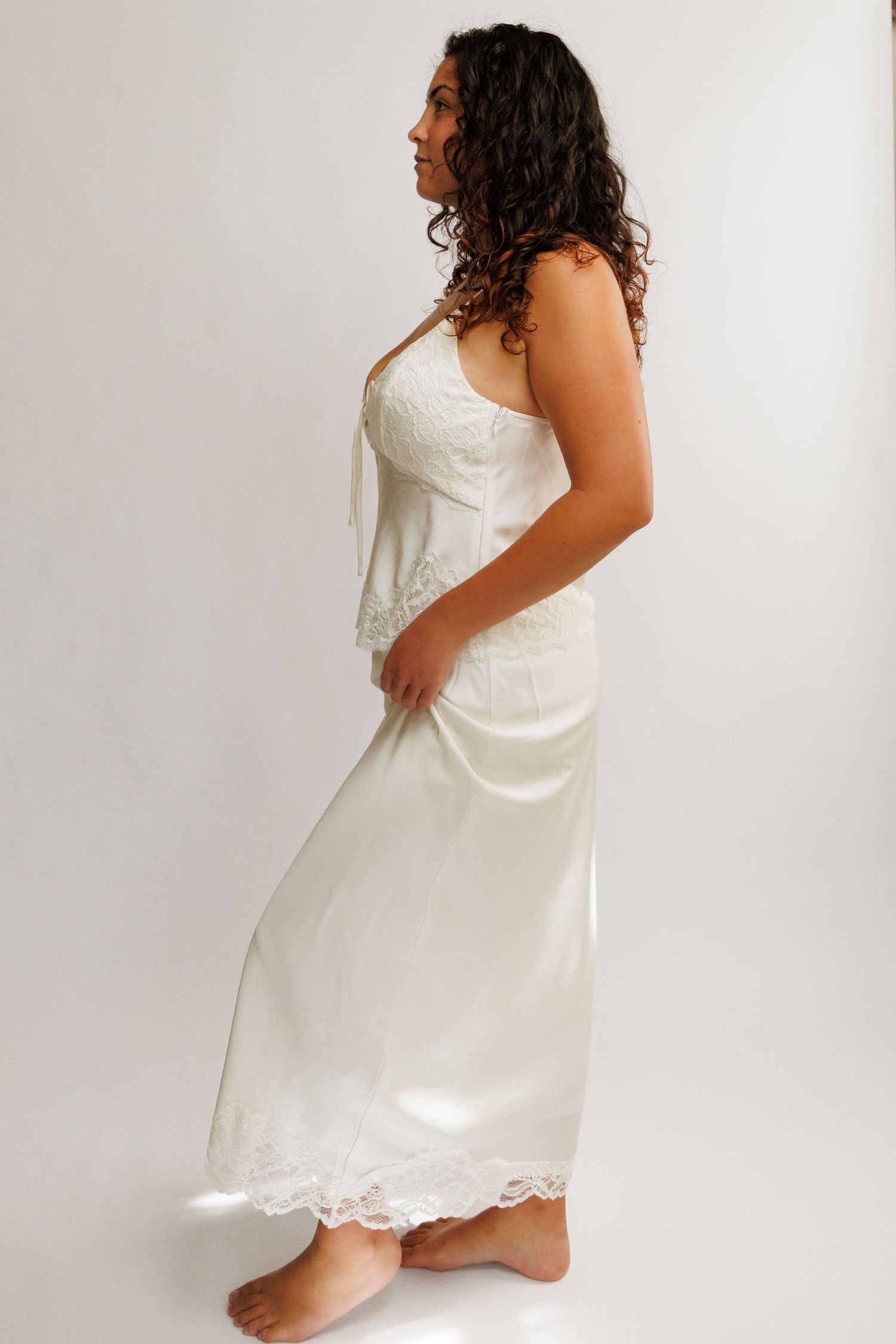Woman in white strapless lace-trimmed bridal gown standing barefoot, side profile view.