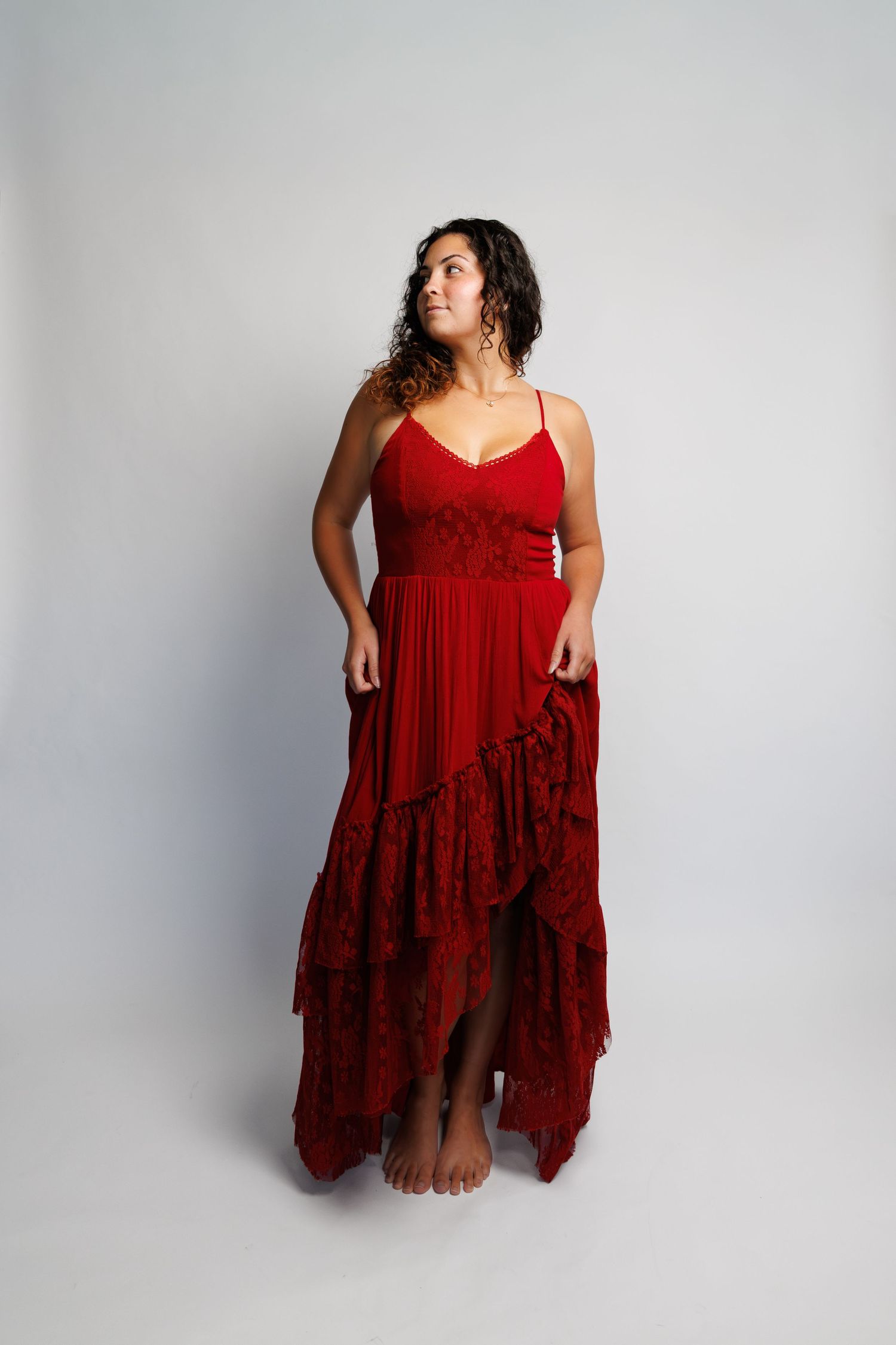 Woman in a flowing red spaghetti strap maxi dress with lace ruffle details, posing barefoot against a white background.