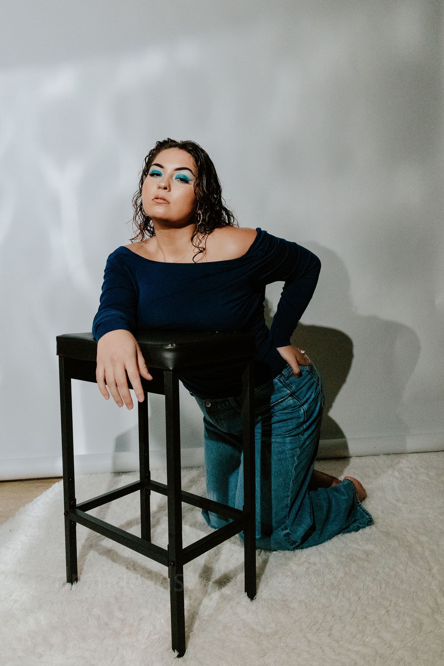 Woman with bold blue eye makeup posing beside a black bar stool in studio setting.
