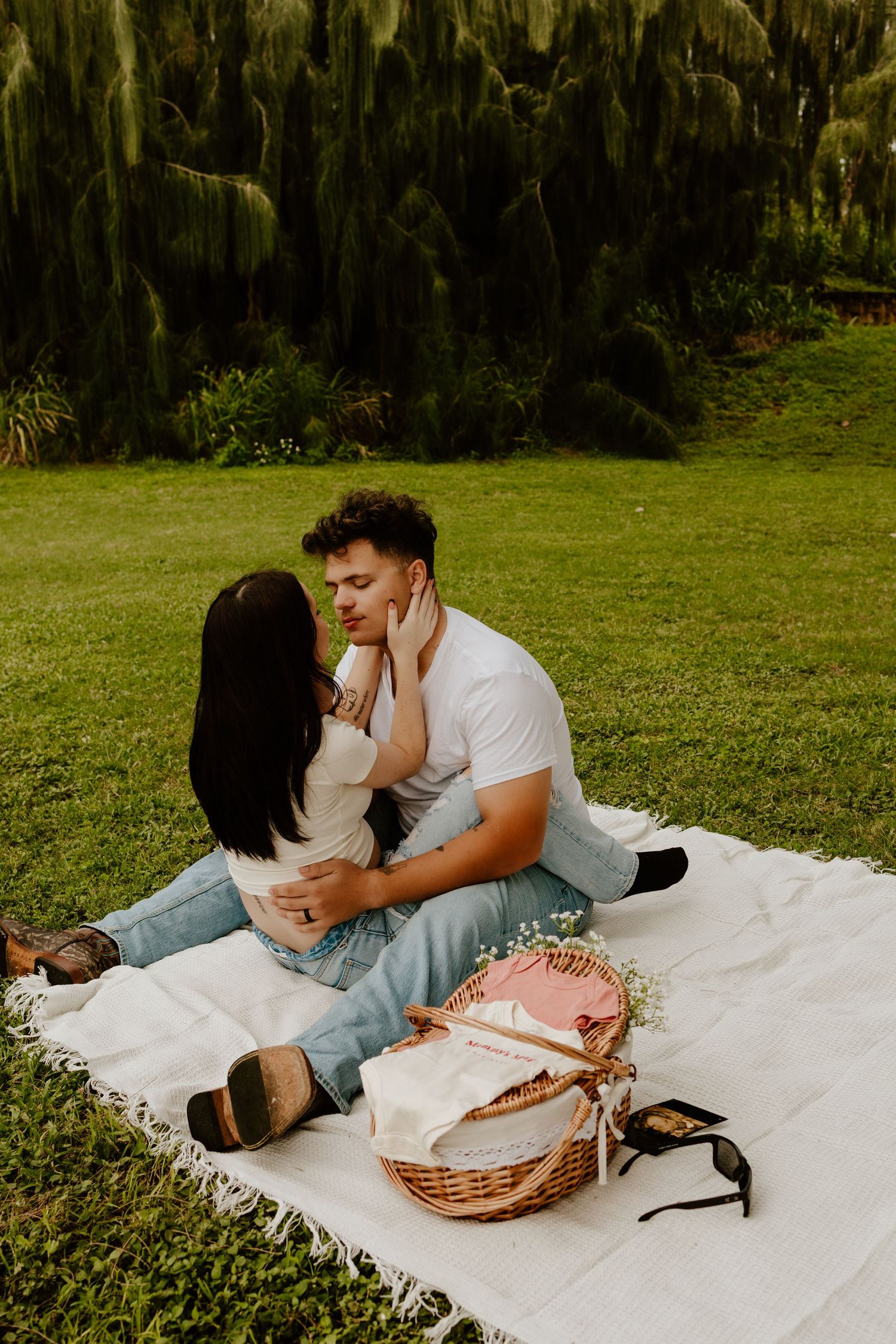Couple sharing a romantic moment on a white blanket during a picnic in a lush green park.