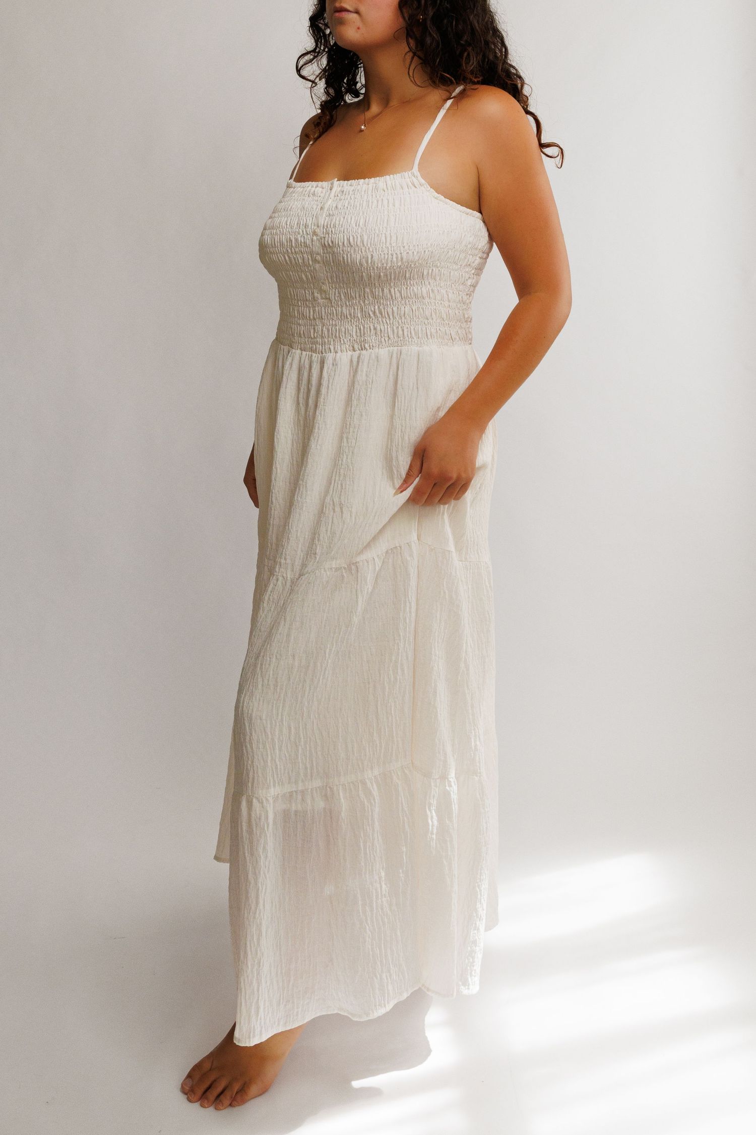 Woman wearing a white smocked bodice maxi dress with spaghetti straps and tiered skirt, barefoot on white background.