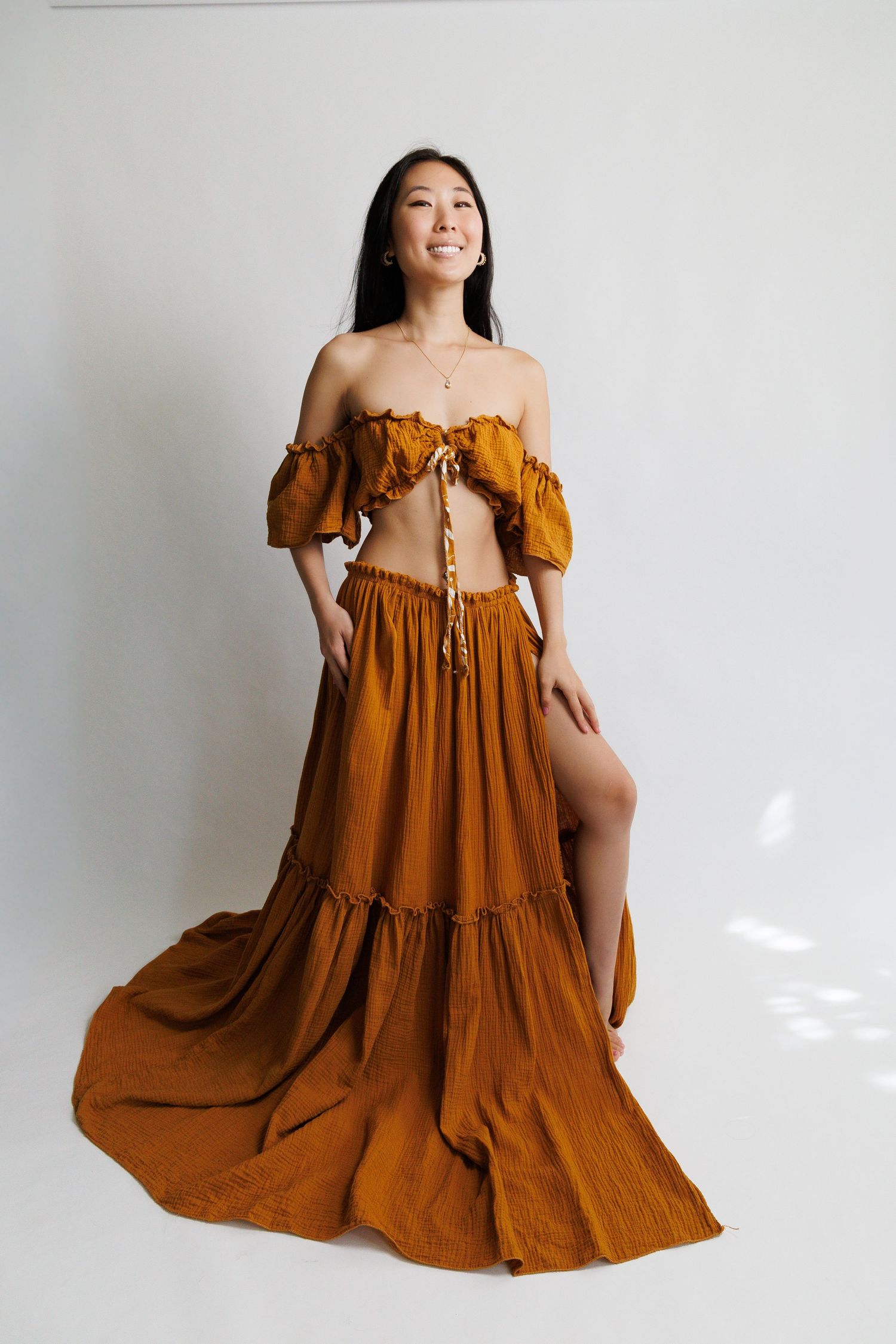 Woman in rust orange off-shoulder crop top and tiered maxi skirt with side slit, posing in studio.