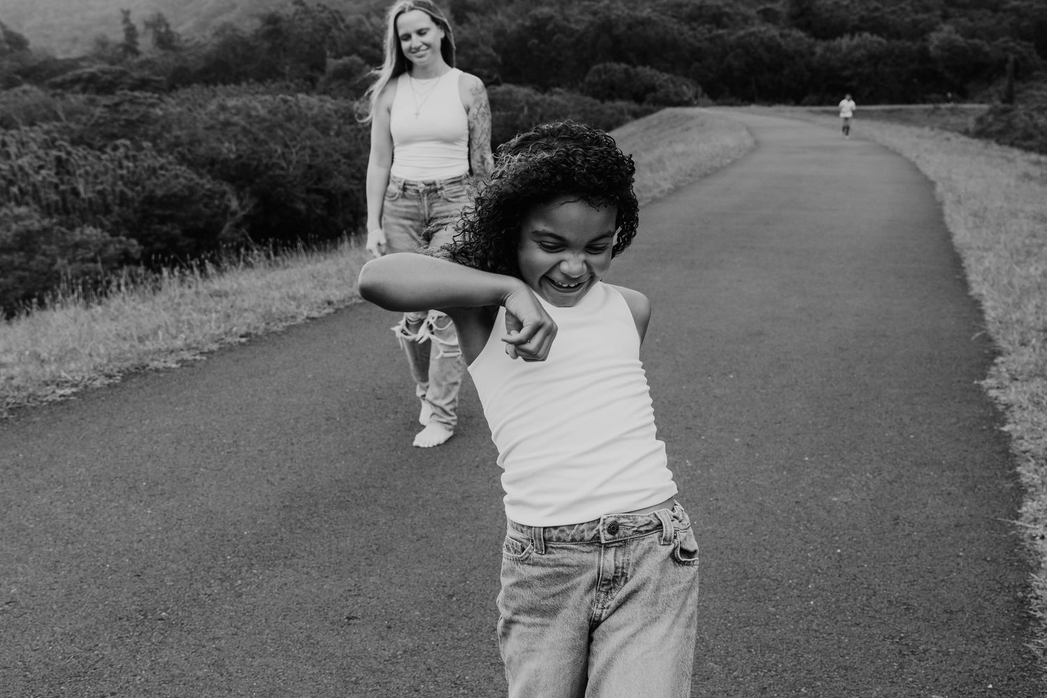 Black and white photo of a girl dancing on a road while a woman watches in the background.