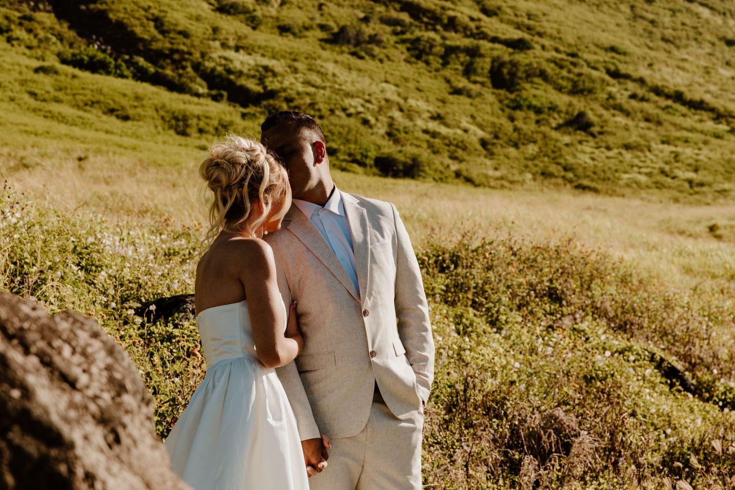 Couple in wedding attire holding hands in sunlit green meadow with rolling hills backdrop.