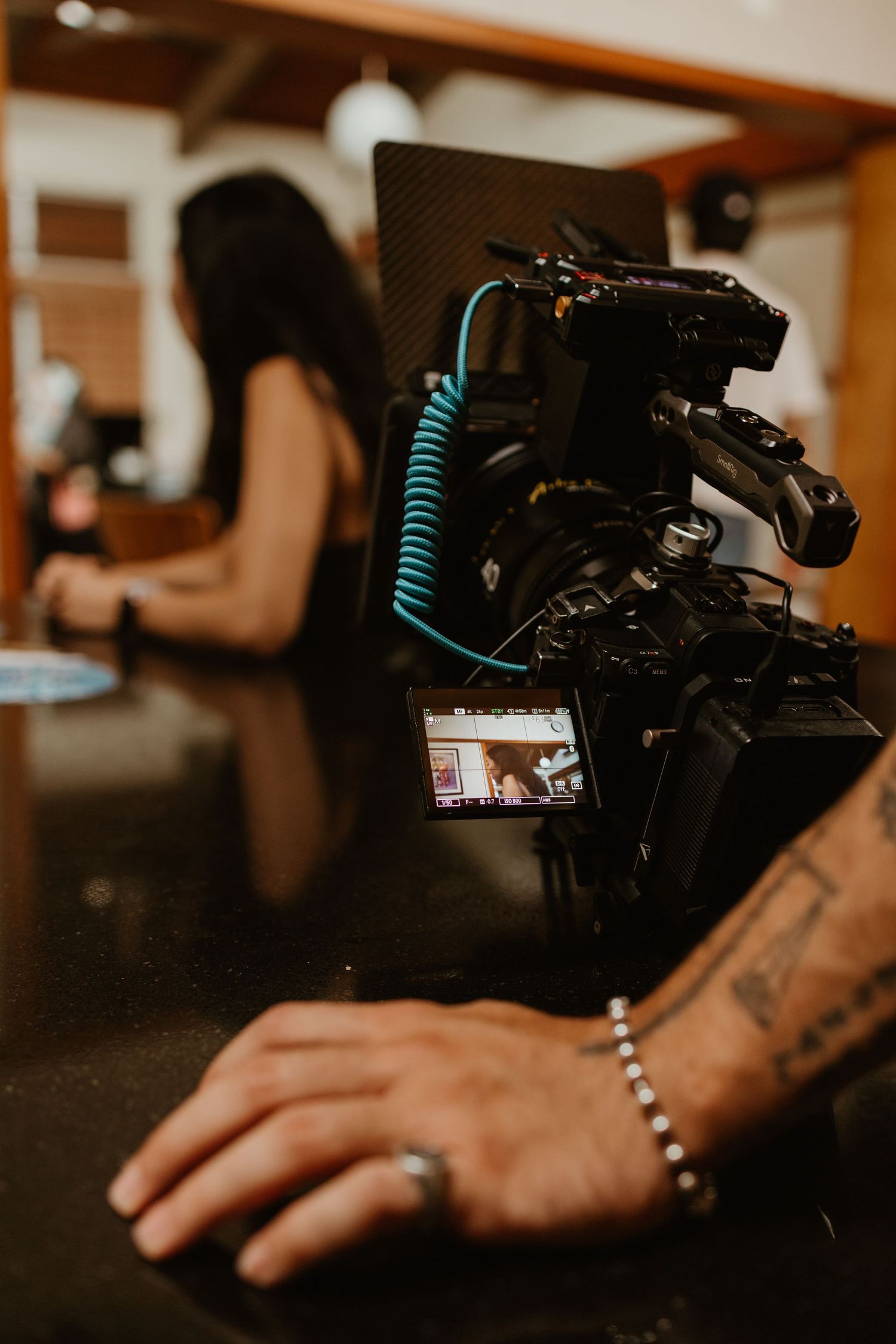 Professional video camera on tripod with teal cable filming a woman in a studio setting, tattooed hand visible in foreground.