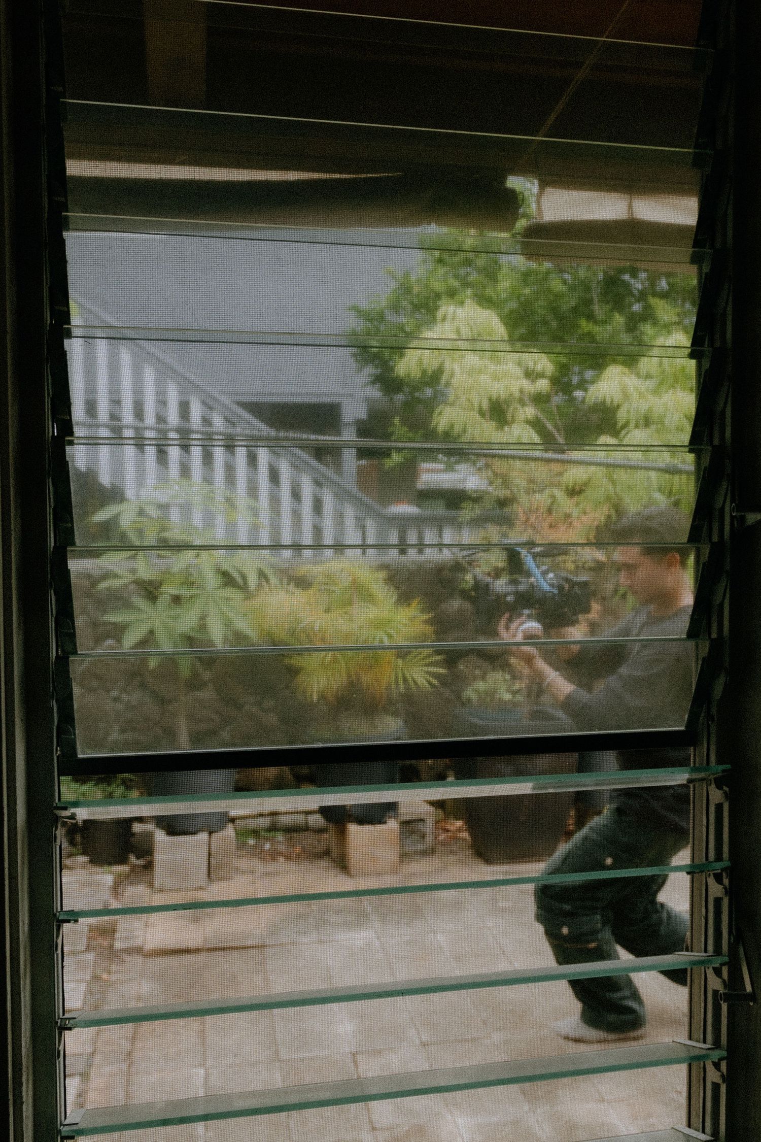 Person cleaning jalousie glass windows with a spray bottle in a lush garden courtyard.
