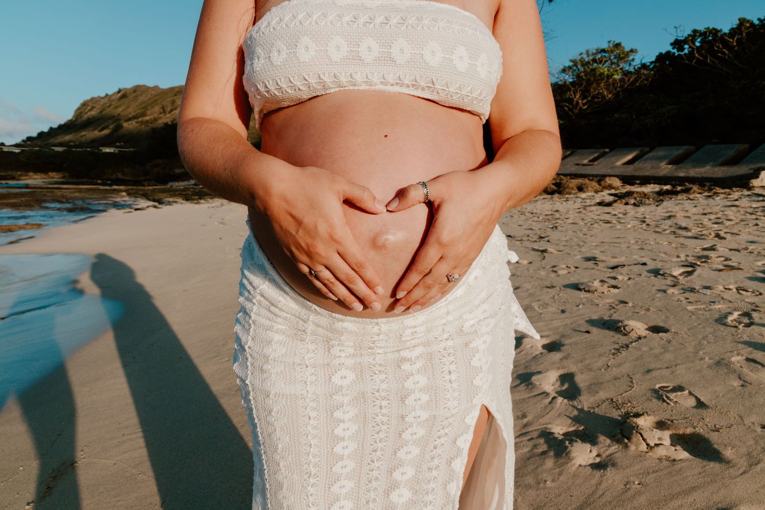 Pregnant woman in white dress making heart shape on baby bump at beach.