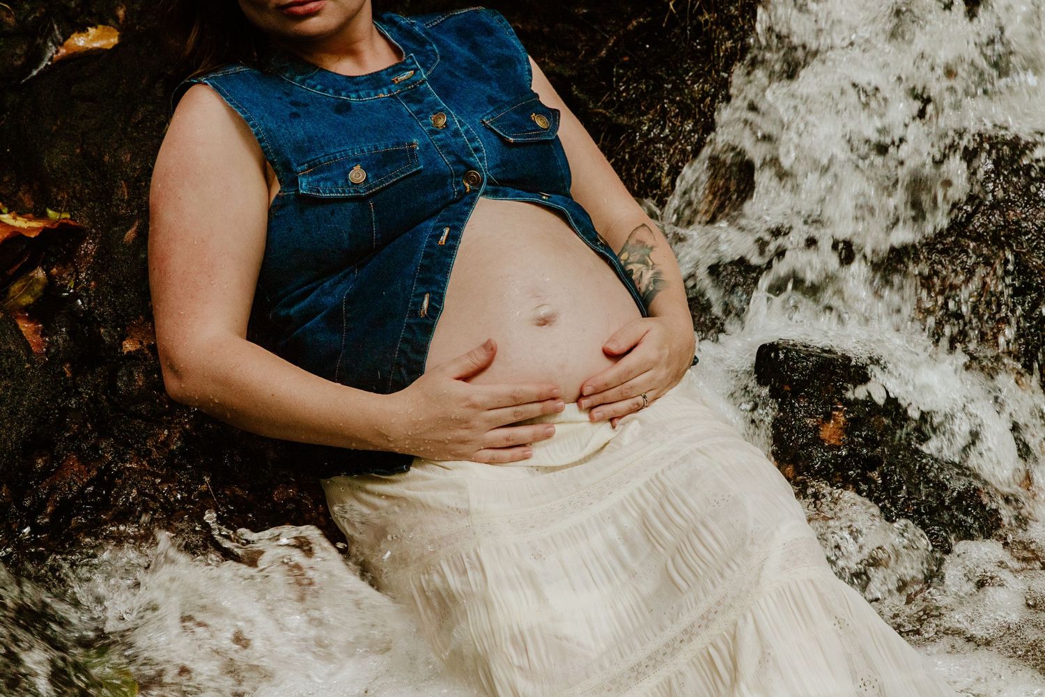 Close-up of pregnant womans bare belly in denim vest and white skirt sitting in flowing waterfall.