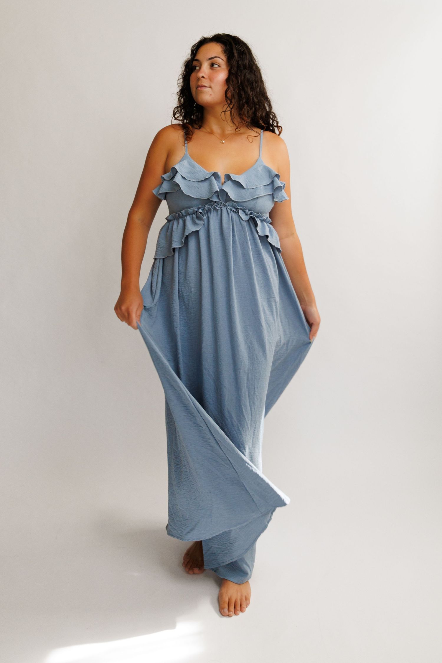 Woman wearing a flowy dusty blue maxi dress with ruffle details and spaghetti straps, posing barefoot.