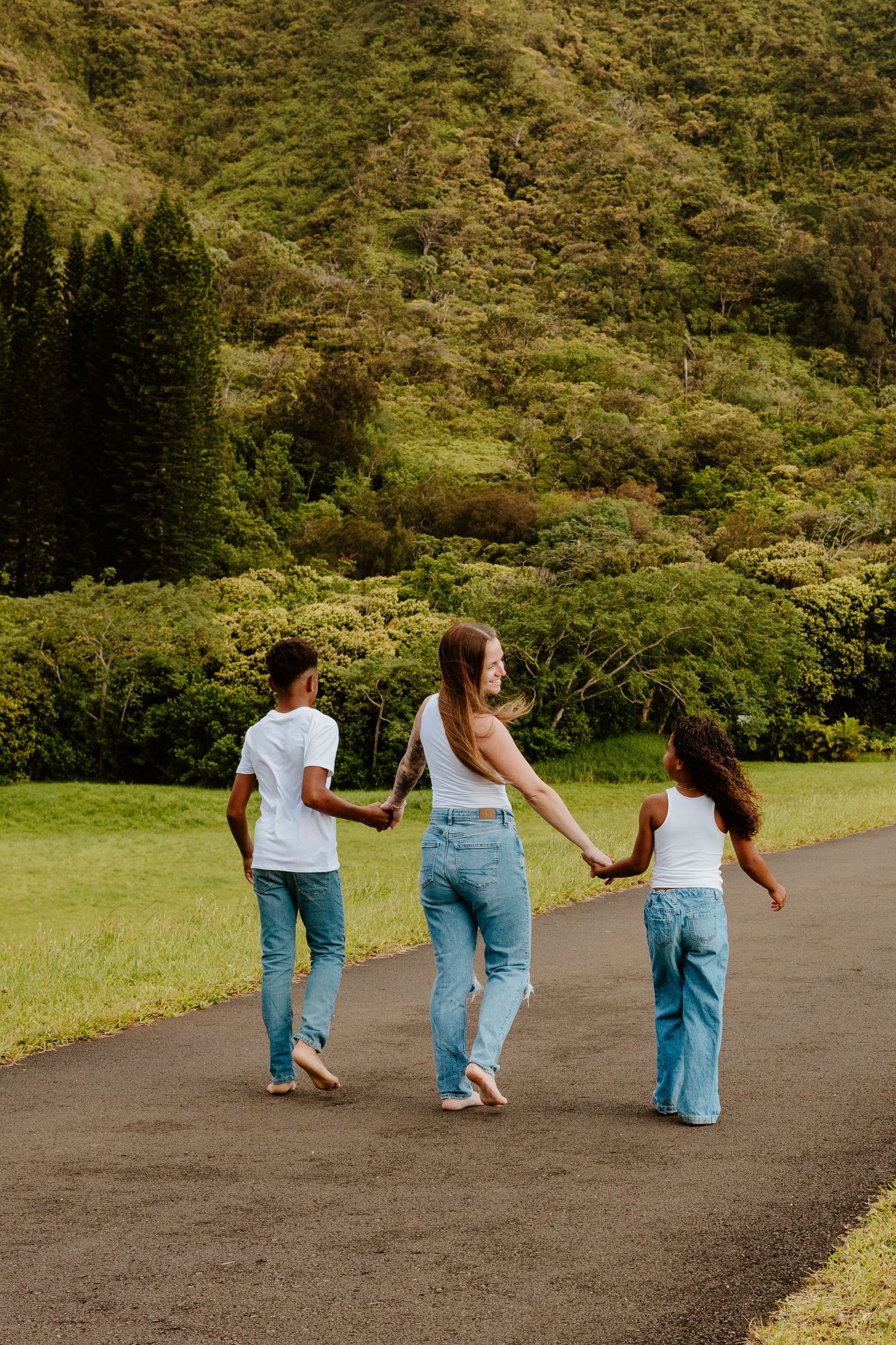 Three people in white tops and jeans walking hand in hand on a road with green hills.