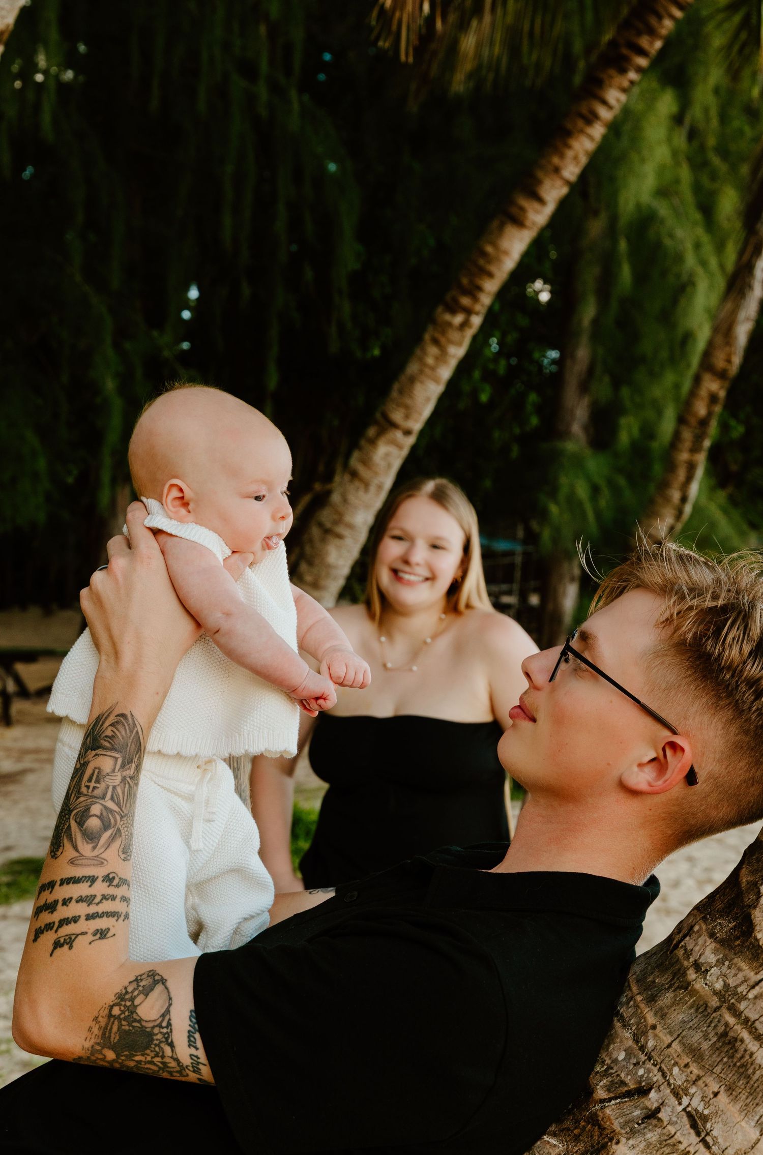 Tattooed father lifting newborn baby while mother smiles under tropical palm trees.