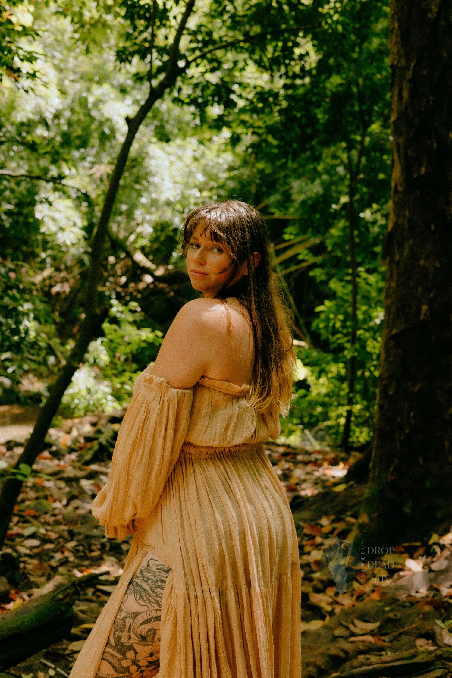 Pregnant woman in golden off-shoulder pleated dress looking back in forest.