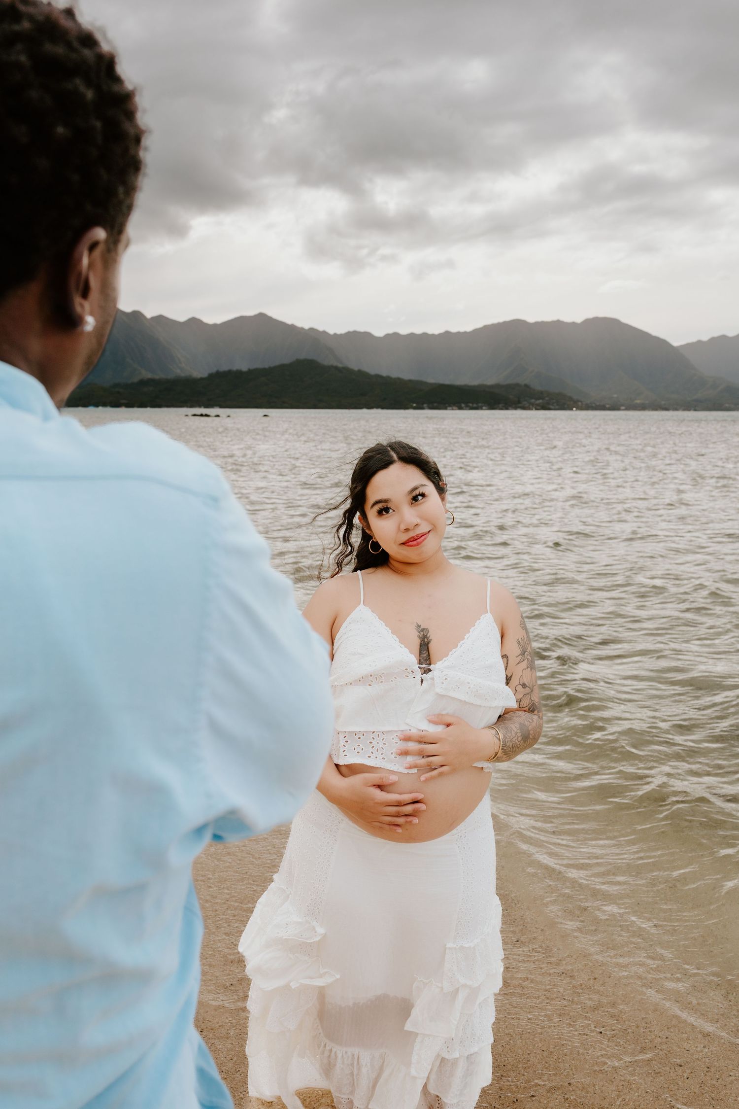 Pregnant tattooed woman in white dress holding belly on beach with partner.