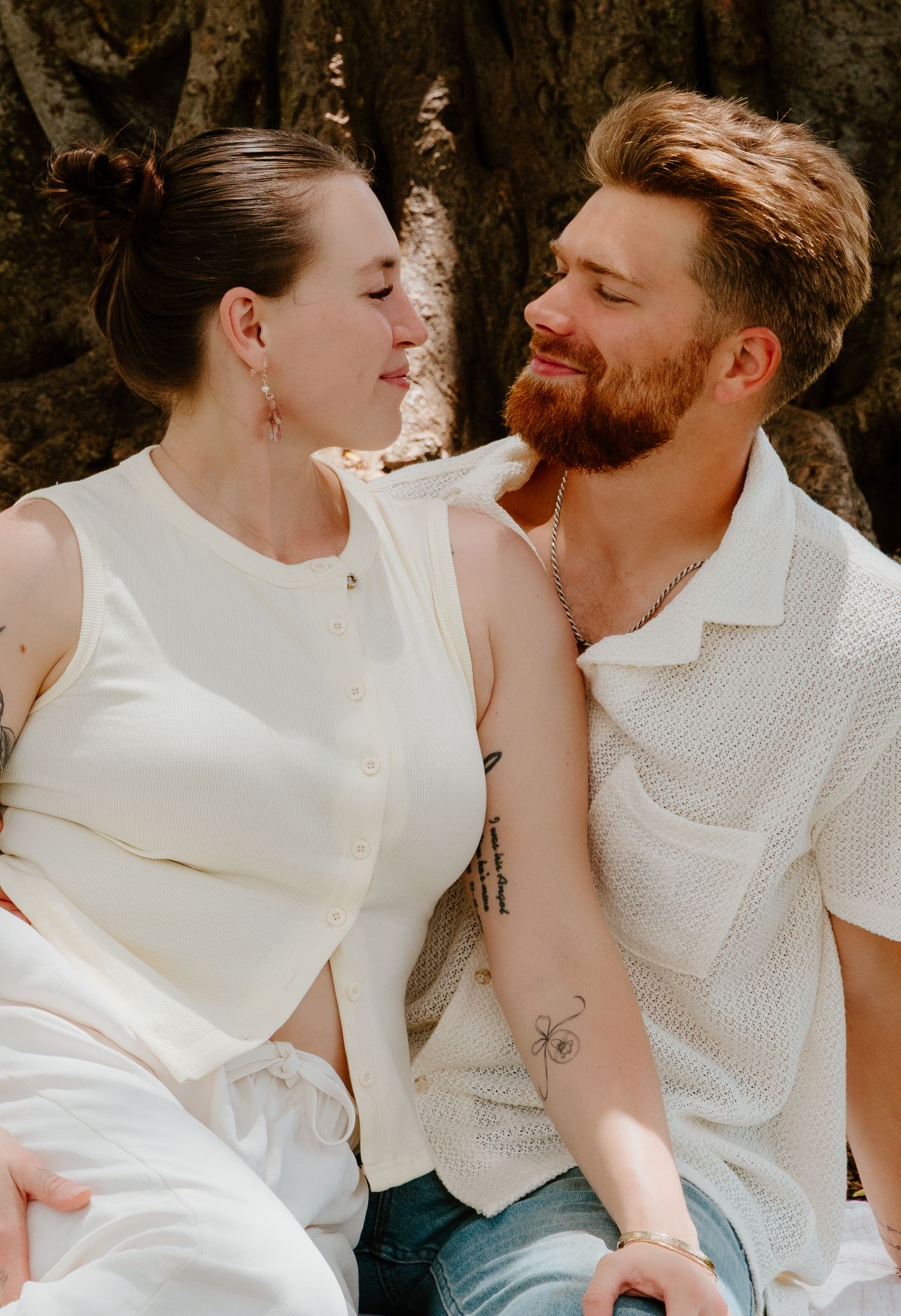 Smiling couple in white outfits sitting together against large tree trunk in sunny park.