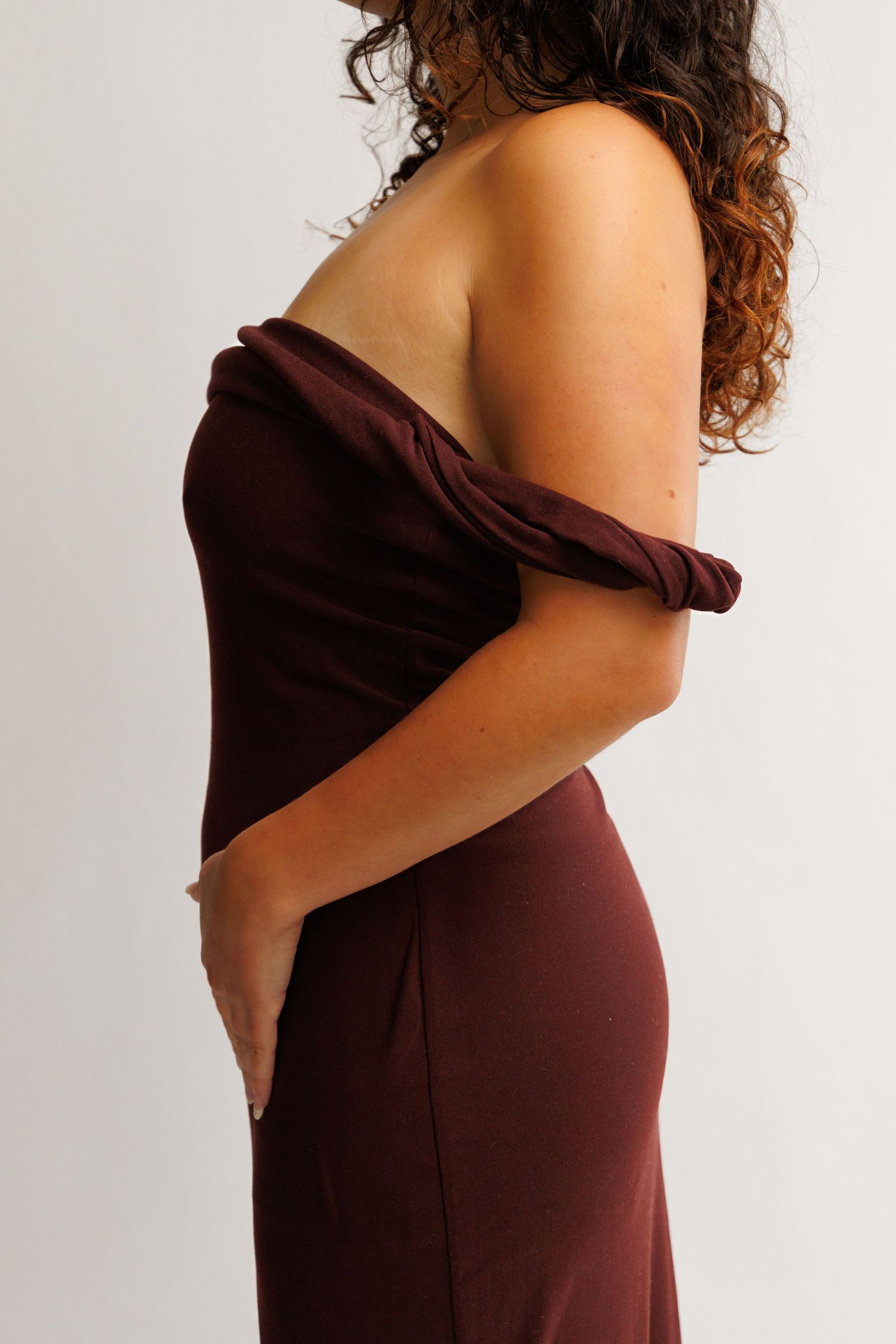 Woman wearing a burgundy off-shoulder fitted gown, shown from the side on a light background.