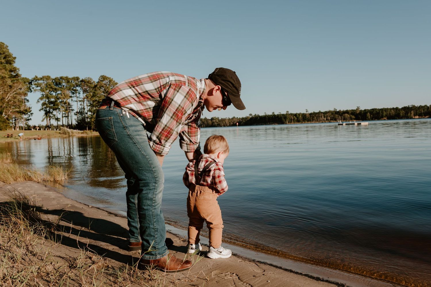 Father in plaid shirt and cap bends down to interact with toddler at the edge of a calm lake at golden hour.