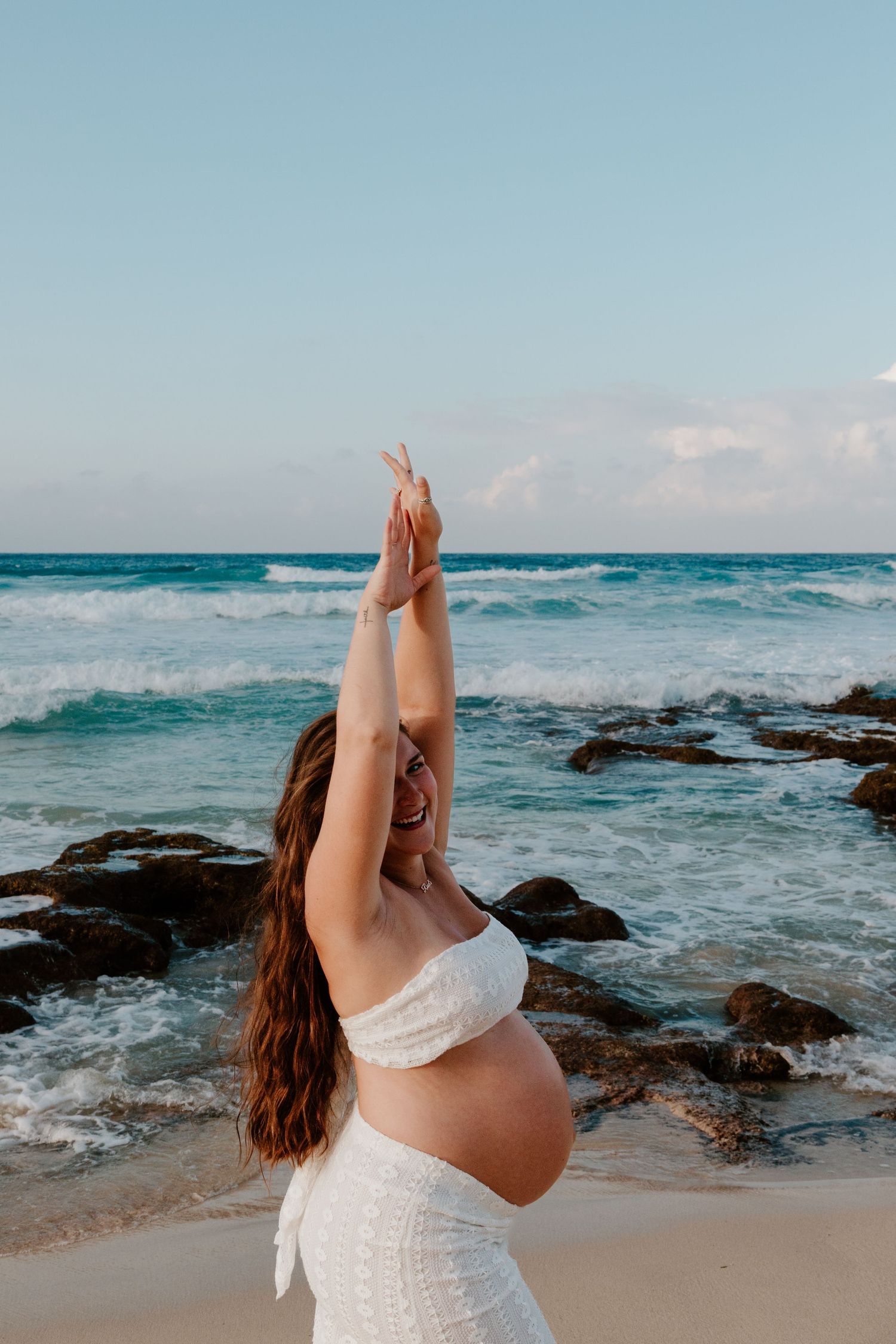 Smiling pregnant woman in white raising arms joyfully at tropical beach.