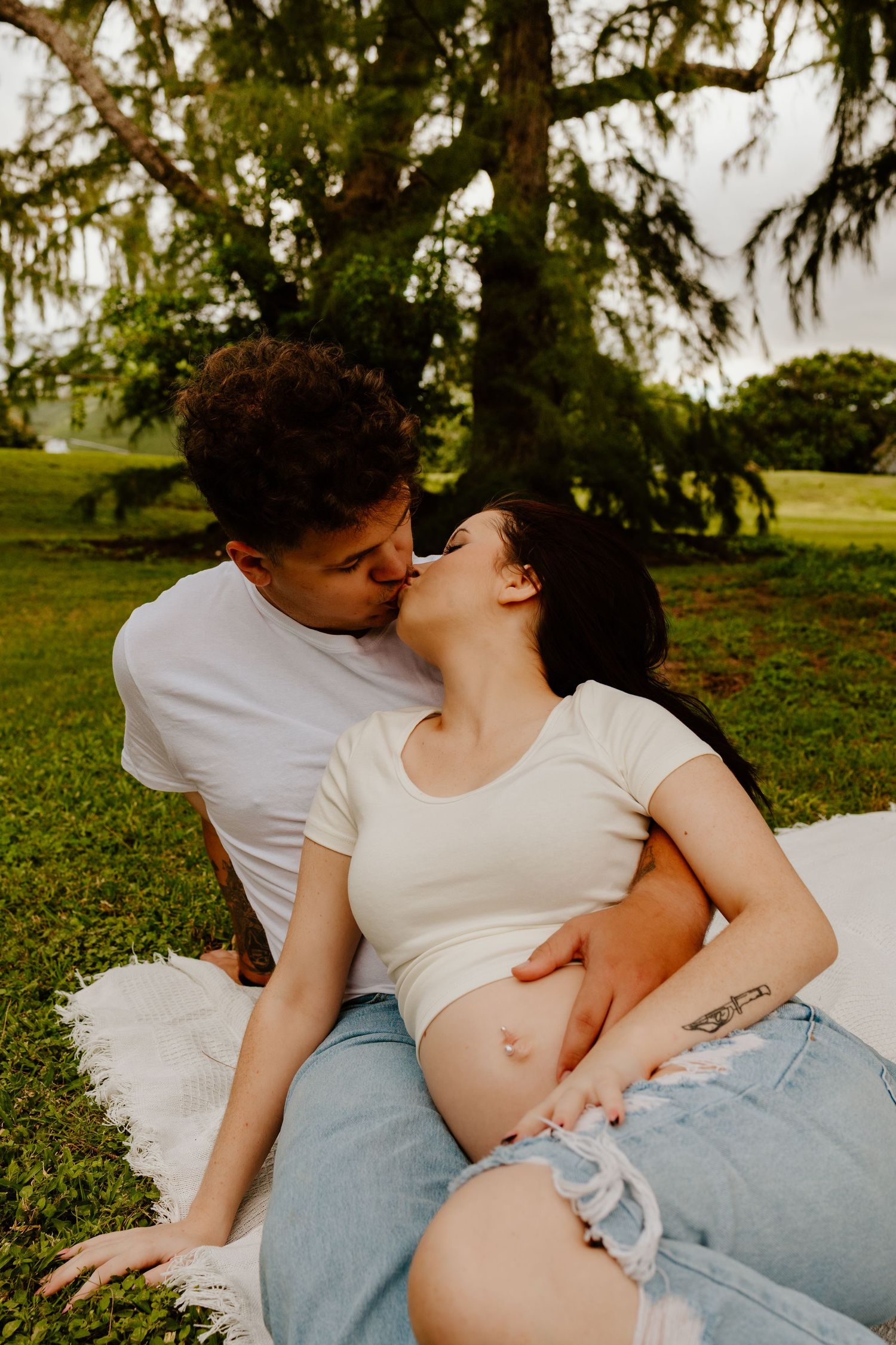 Couple sharing tender kiss on picnic blanket outdoors, pregnant woman in white crop top.