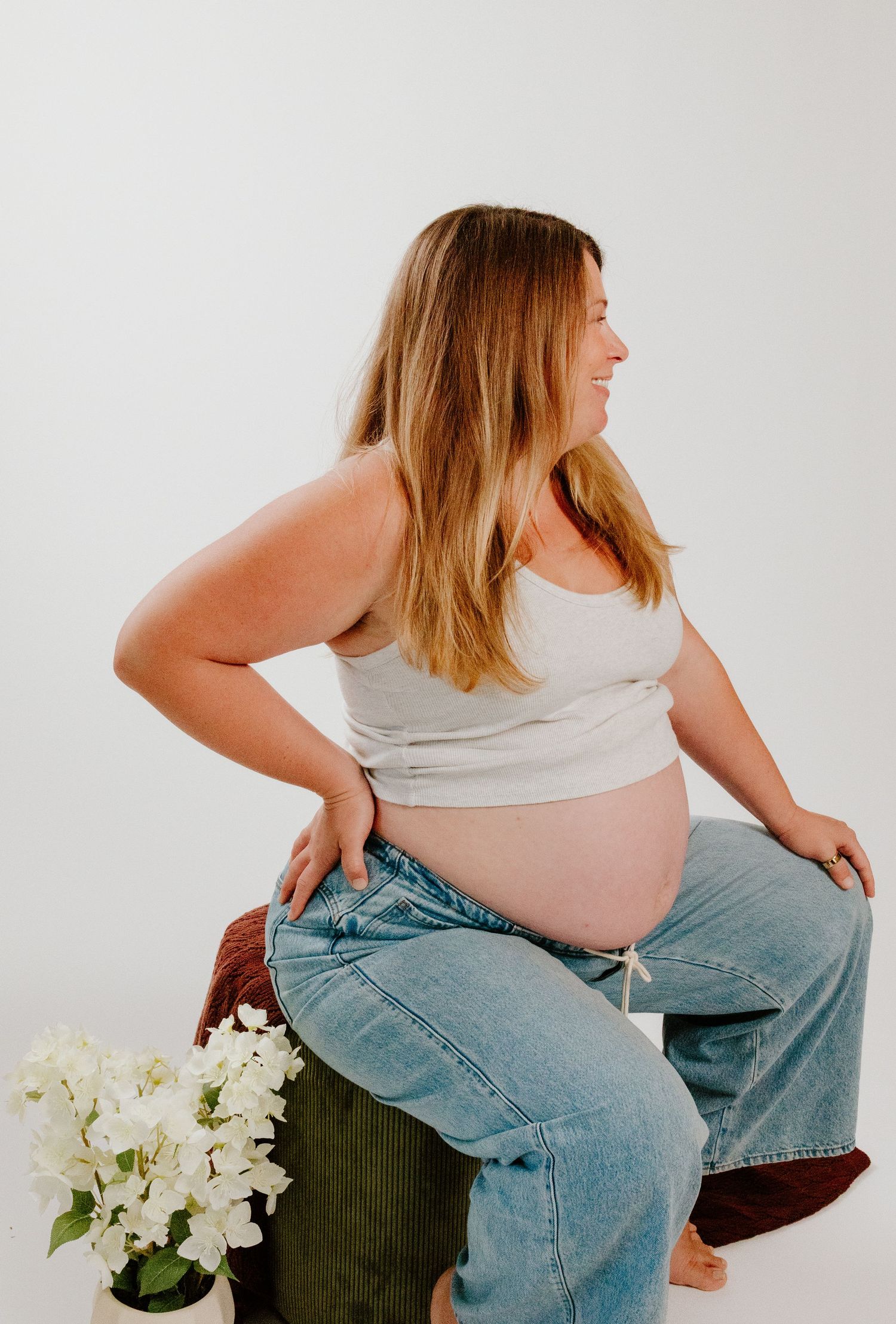Smiling pregnant blonde woman in crop top and jeans sitting during maternity shoot.