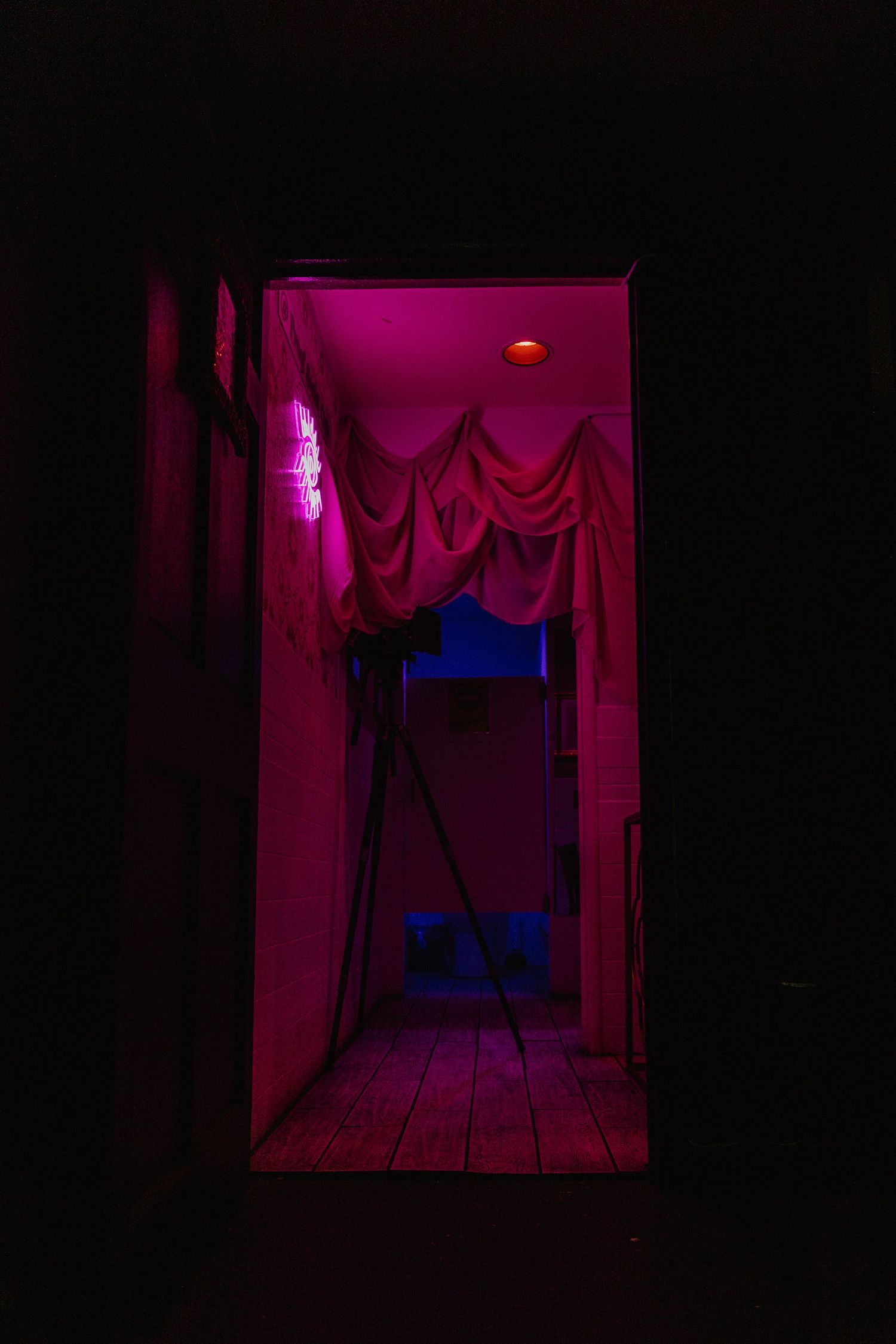 Dark hallway illuminated by pink and blue neon lights with draped fabric curtains and wooden floor.