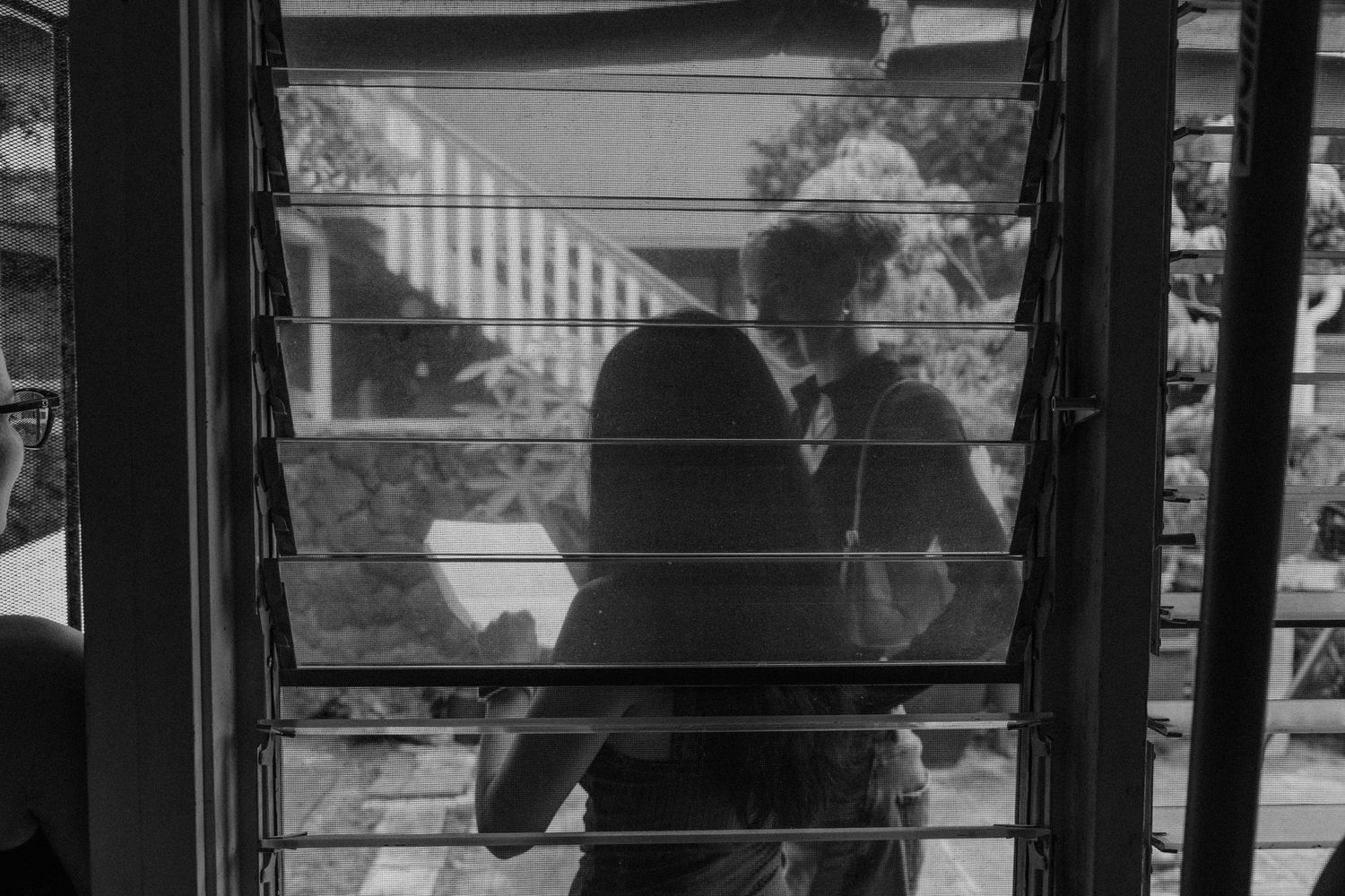 Black and white photo of a couple kissing on a porch, viewed through window blinds.