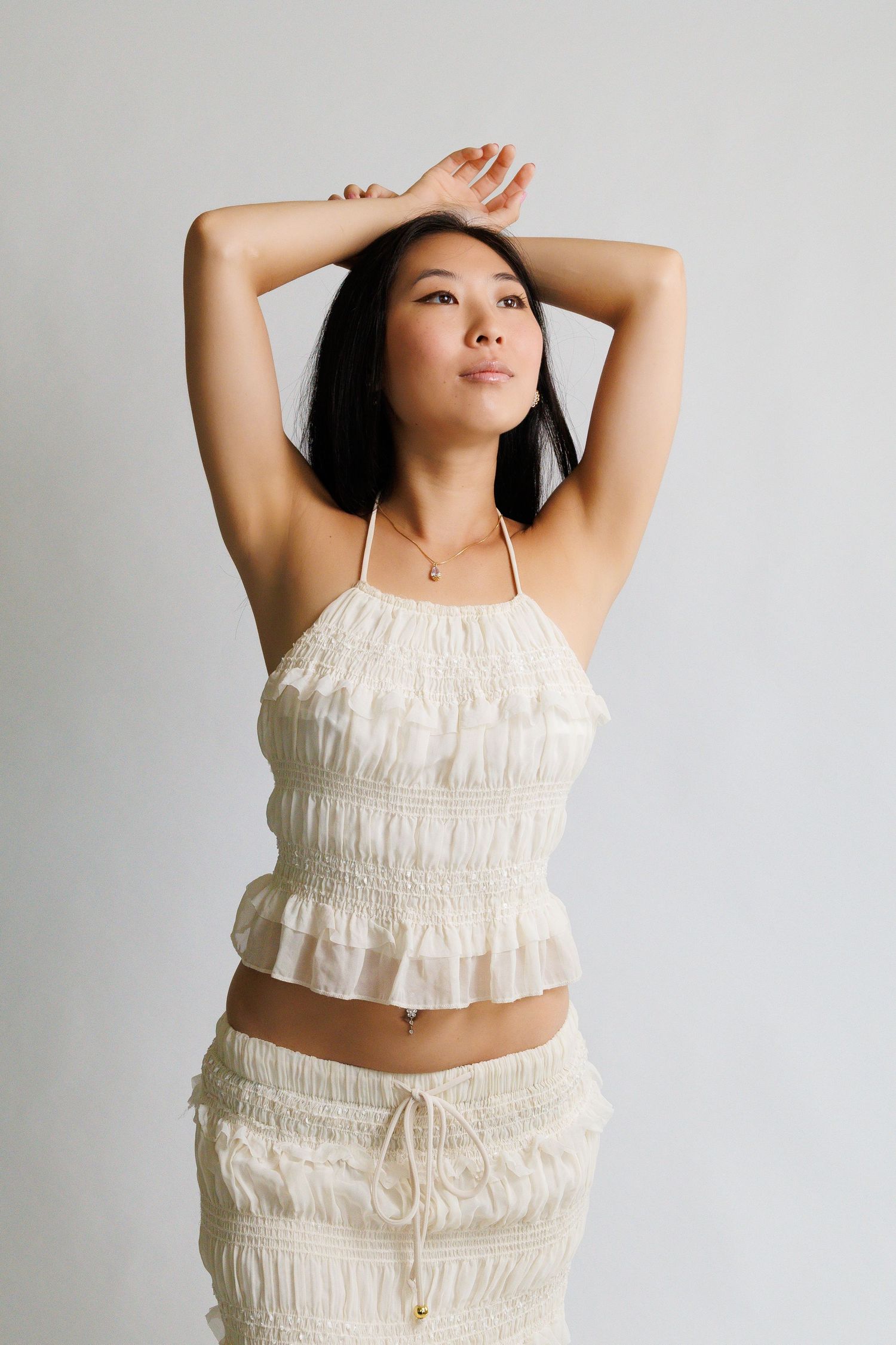Woman in cream ruffled halter top and matching skirt posing with arms raised above head against white background.