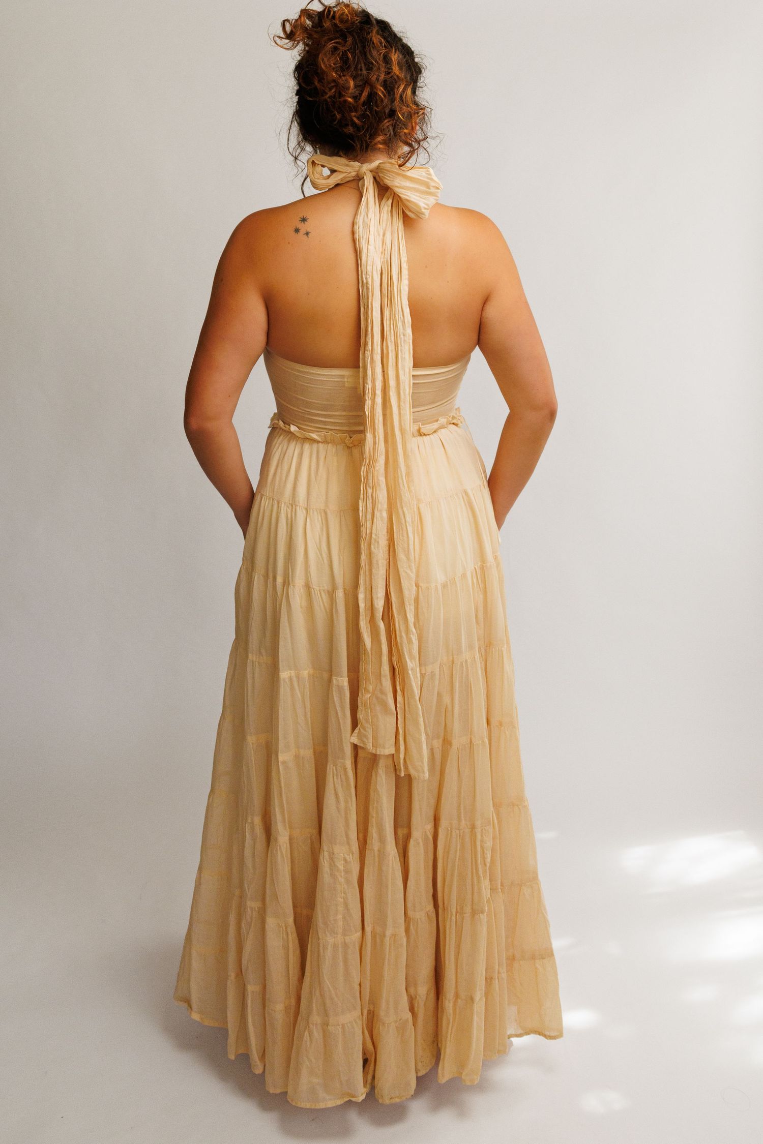Woman wearing a flowing gold halter maxi dress with tiered skirt, viewed from behind, showing open back design.