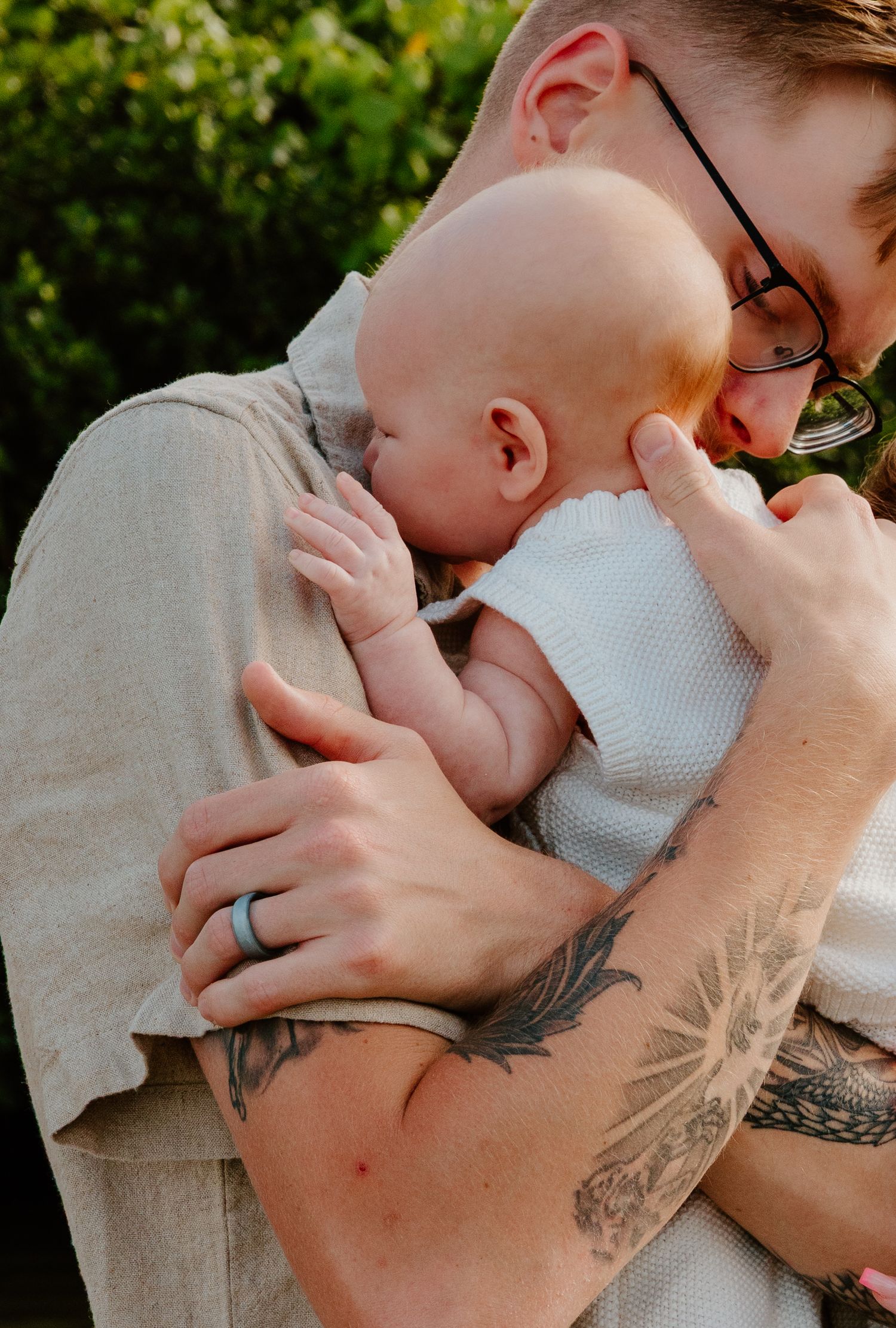 Tattooed father wearing glasses and wedding ring gently holding sleeping newborn baby.