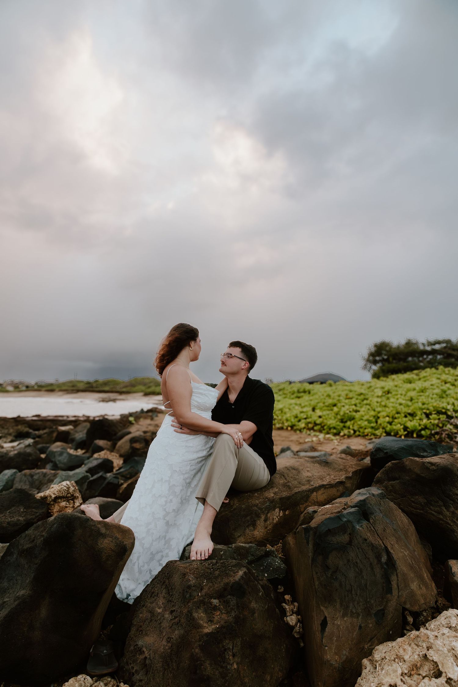 A couple sits on rocky shore gazing at each other in wedding attire under a dramatic cloudy sky.