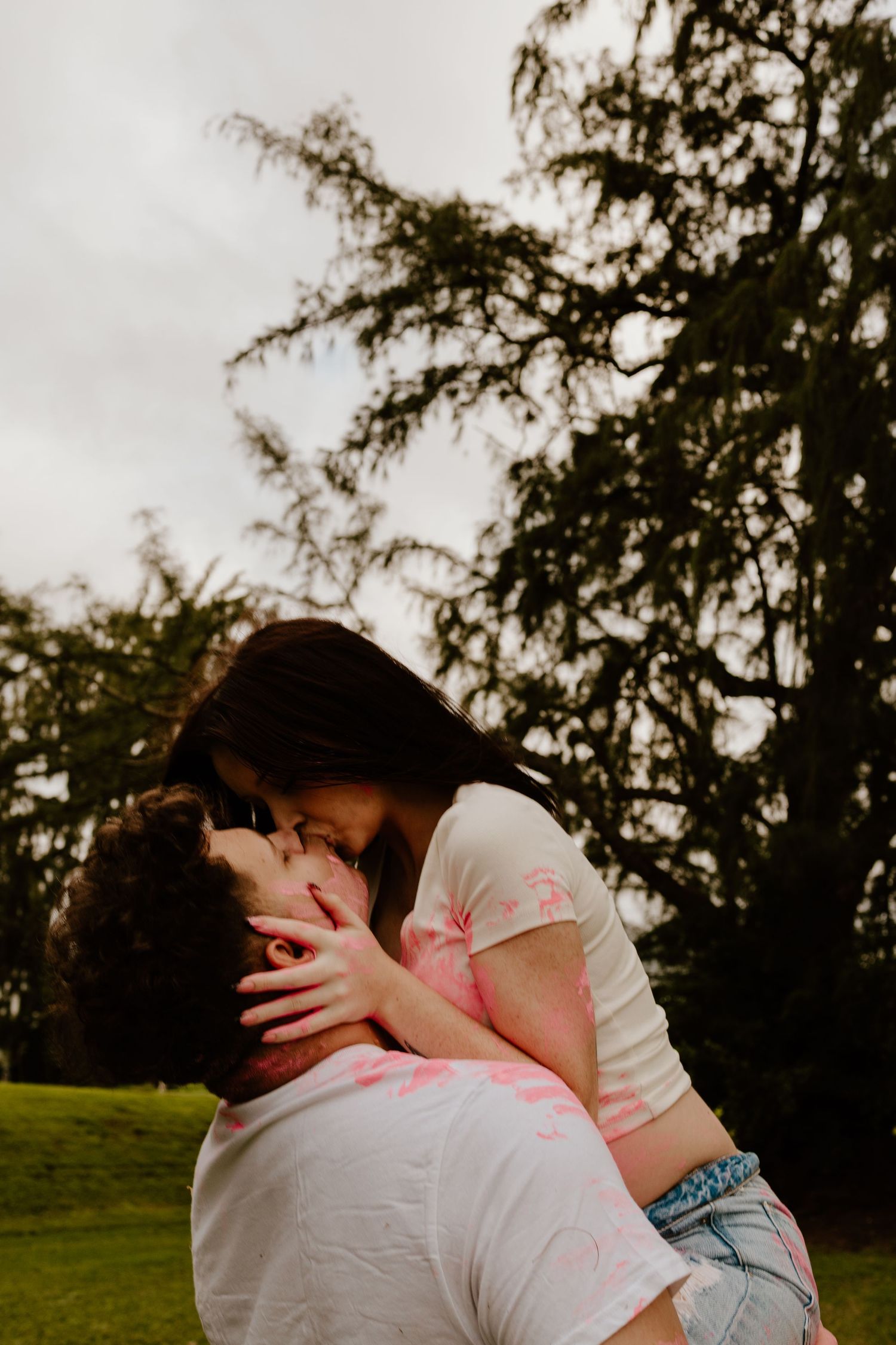 A couple shares a passionate kiss outdoors with dramatic evergreen trees in the background.