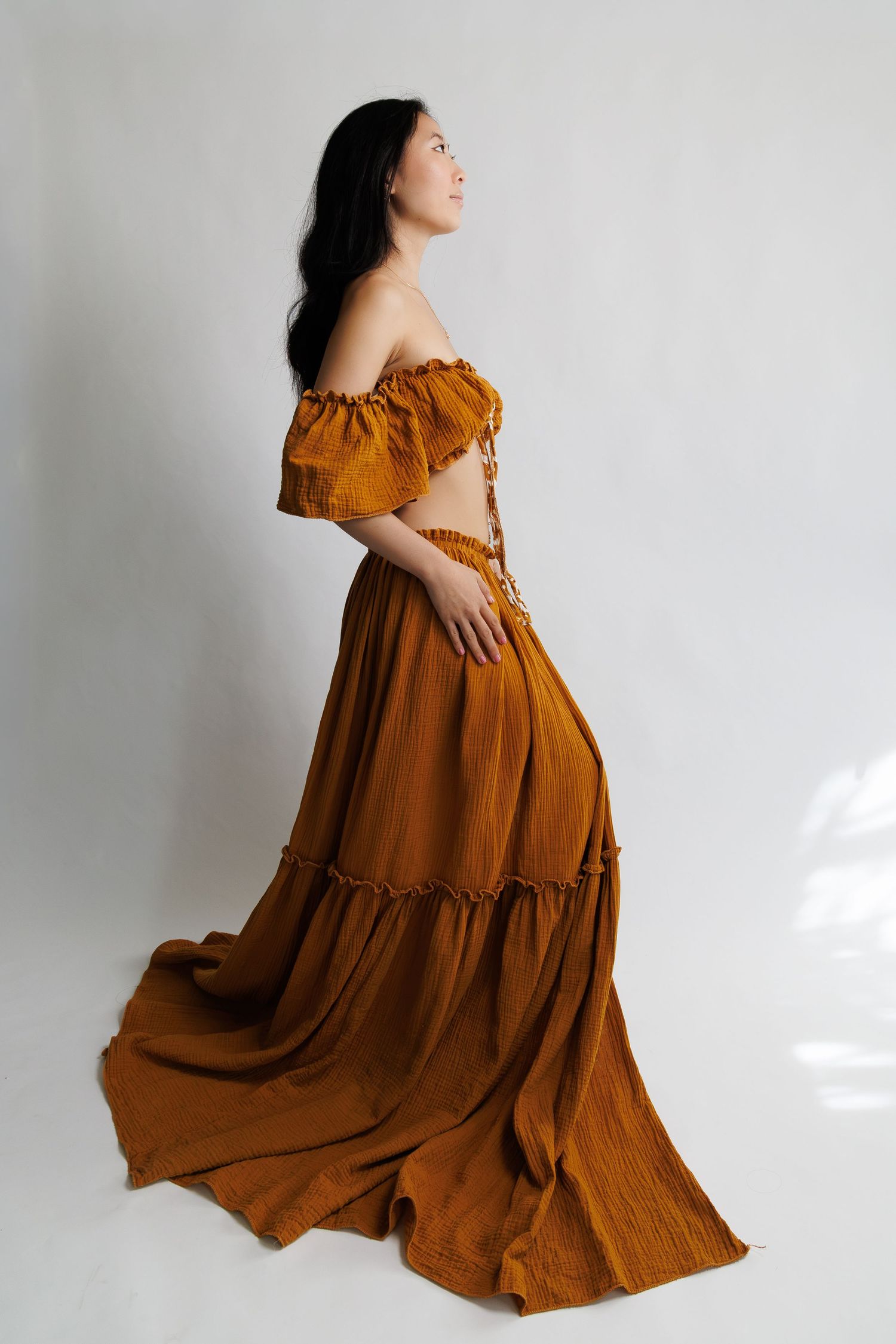 Woman in rust orange off-shoulder tiered maxi dress with flowing skirt, posing in profile view.