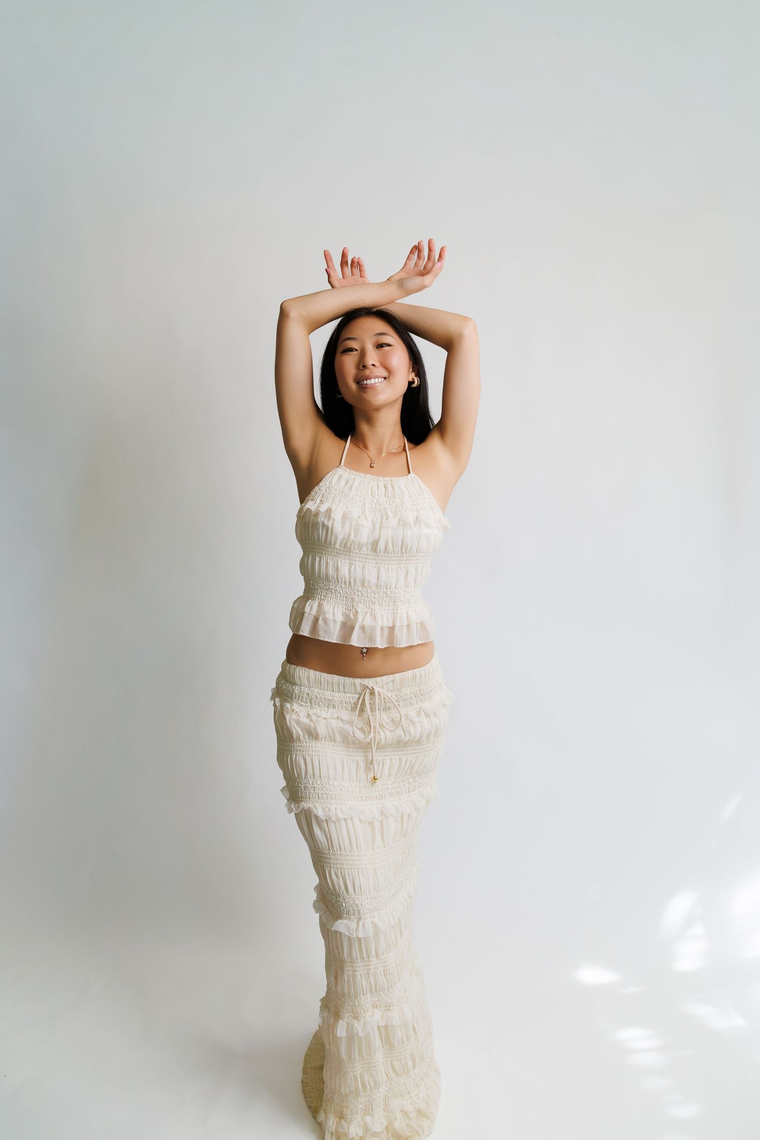 Woman in white ruffled crop top and maxi skirt posing with arms raised overhead, smiling against white background.