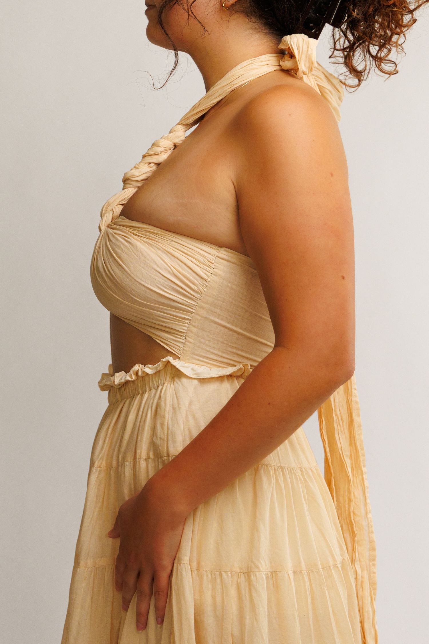 Woman wearing a cream ruched halter maxi dress with cutout waist and tiered skirt, side profile view.