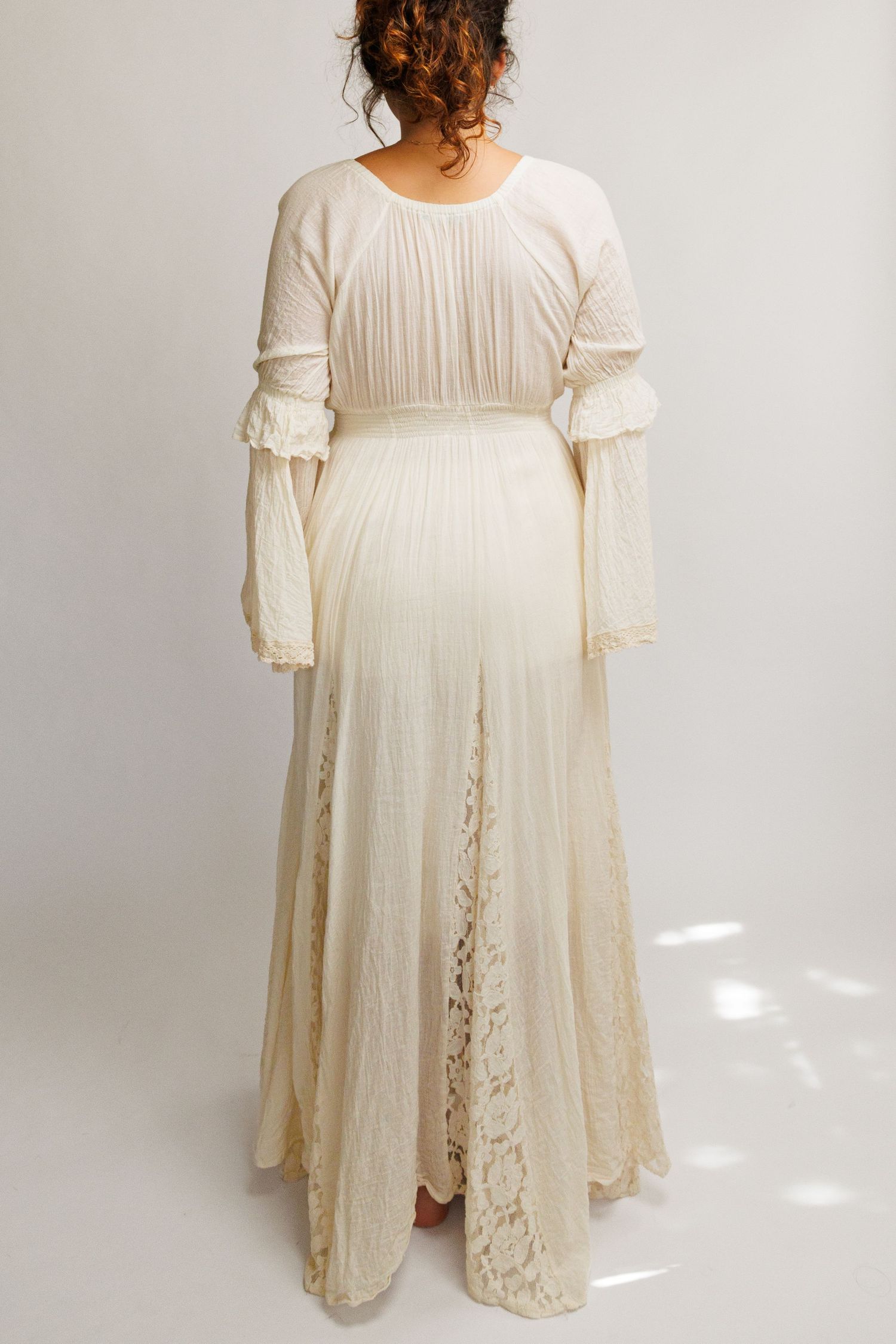Woman wearing an ivory cream boho maxi dress with lace skirt, pleated bodice, and bell sleeves, shown from behind.