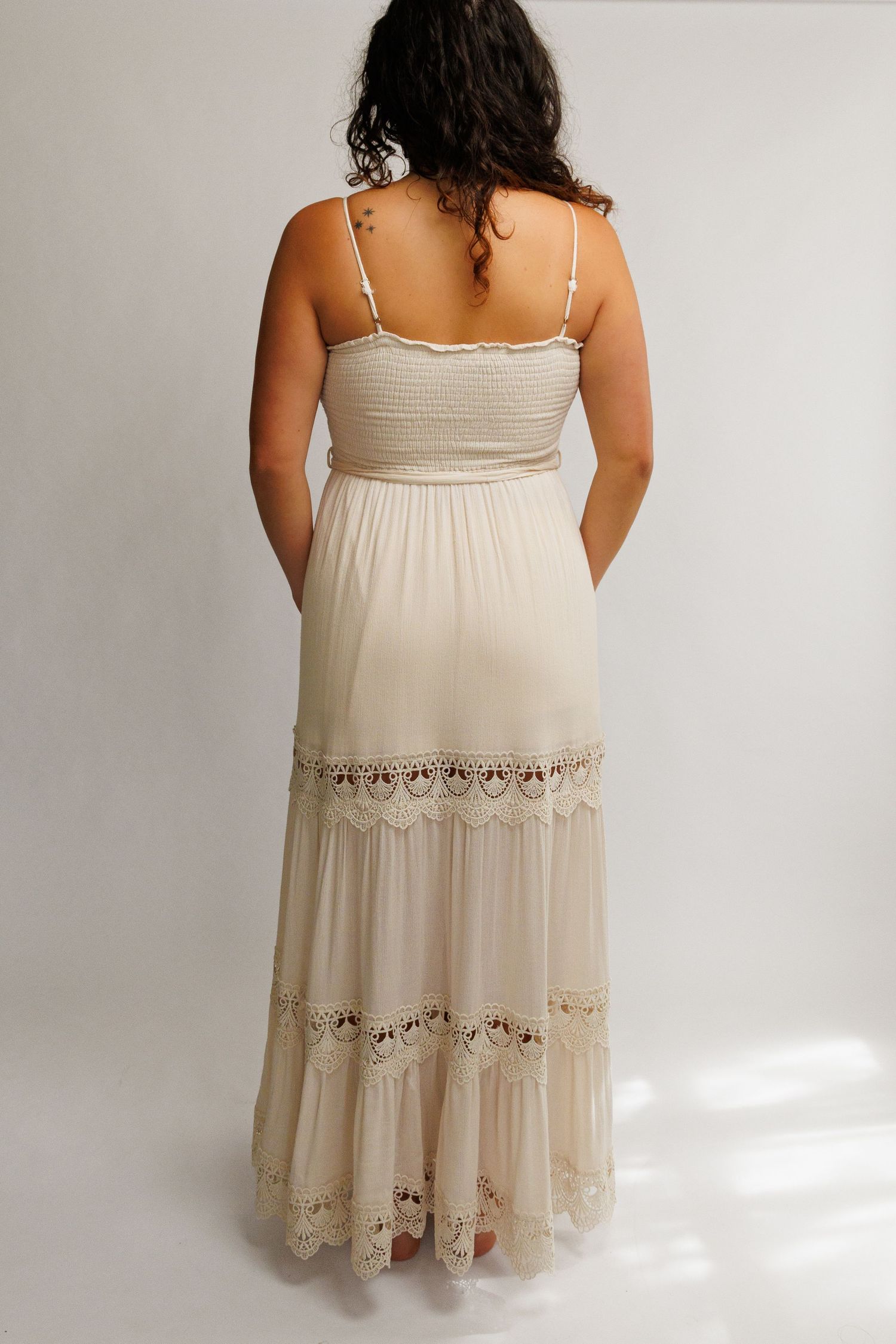 Back view of a woman wearing a cream boho maxi dress with lace trim, smocked bodice, and spaghetti straps.
