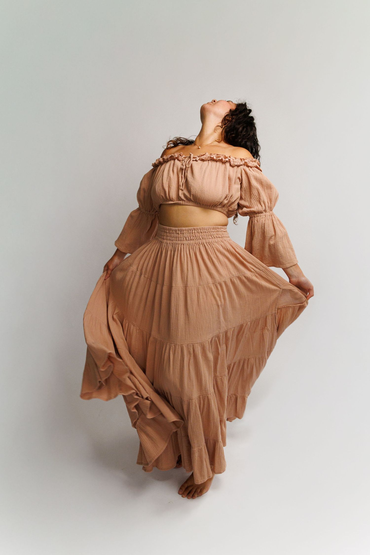 Woman in flowing terracotta off-shoulder crop top and maxi skirt twirling gracefully against white background.