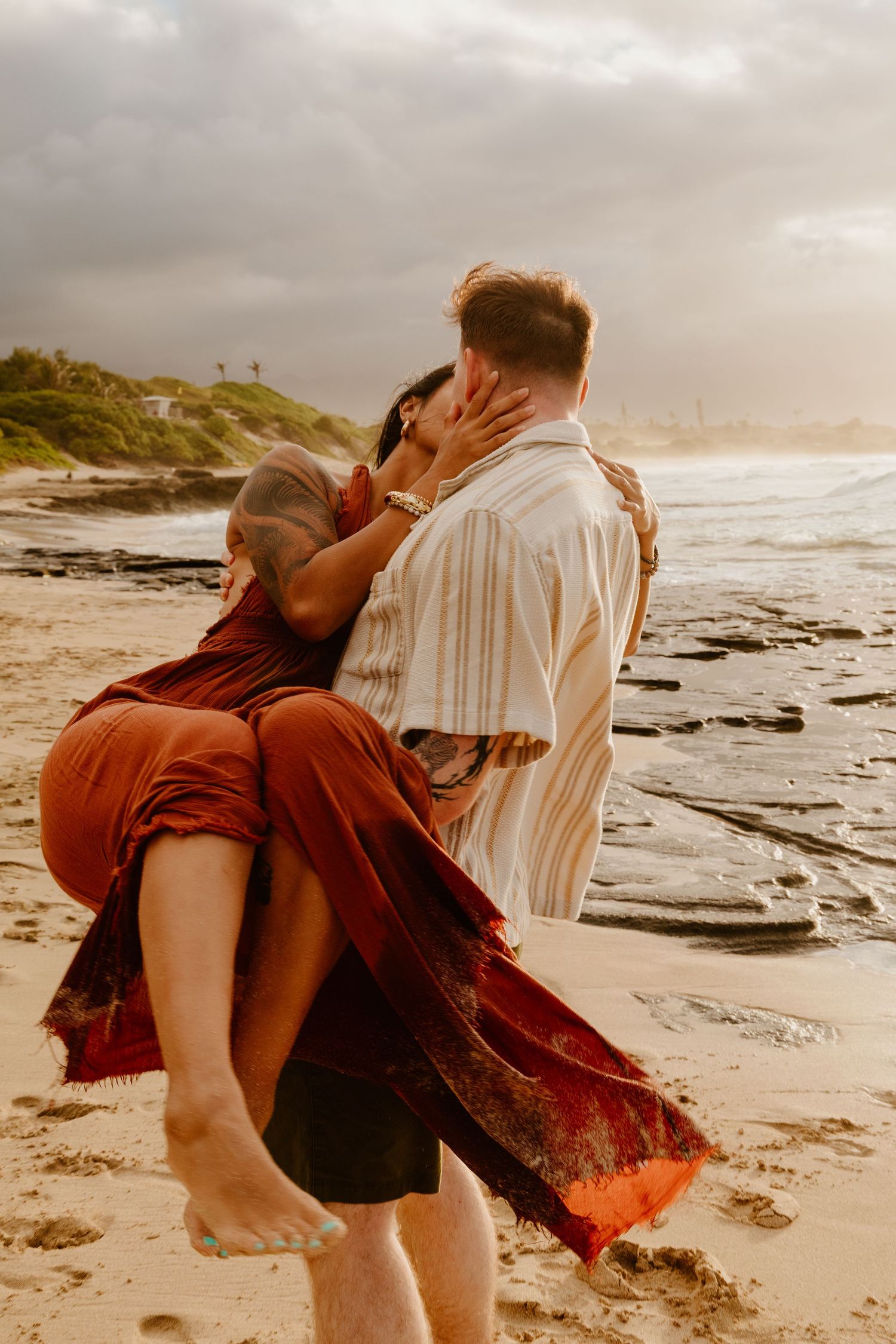 Couple sharing a romantic kiss on a sandy beach at sunset, woman in flowing rust dress being lifted by man in striped shirt.