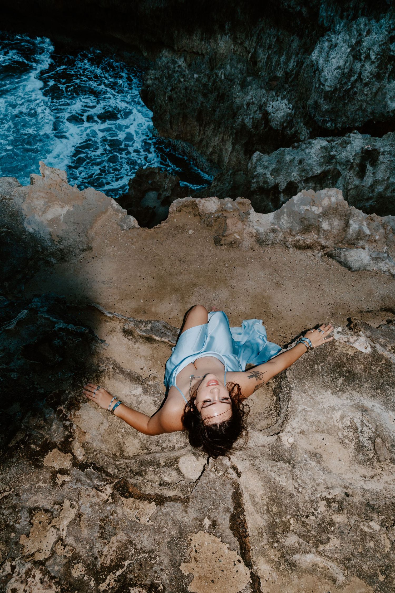 Woman in blue dress lying on rocky coastal cliffs with turquoise ocean waves below.