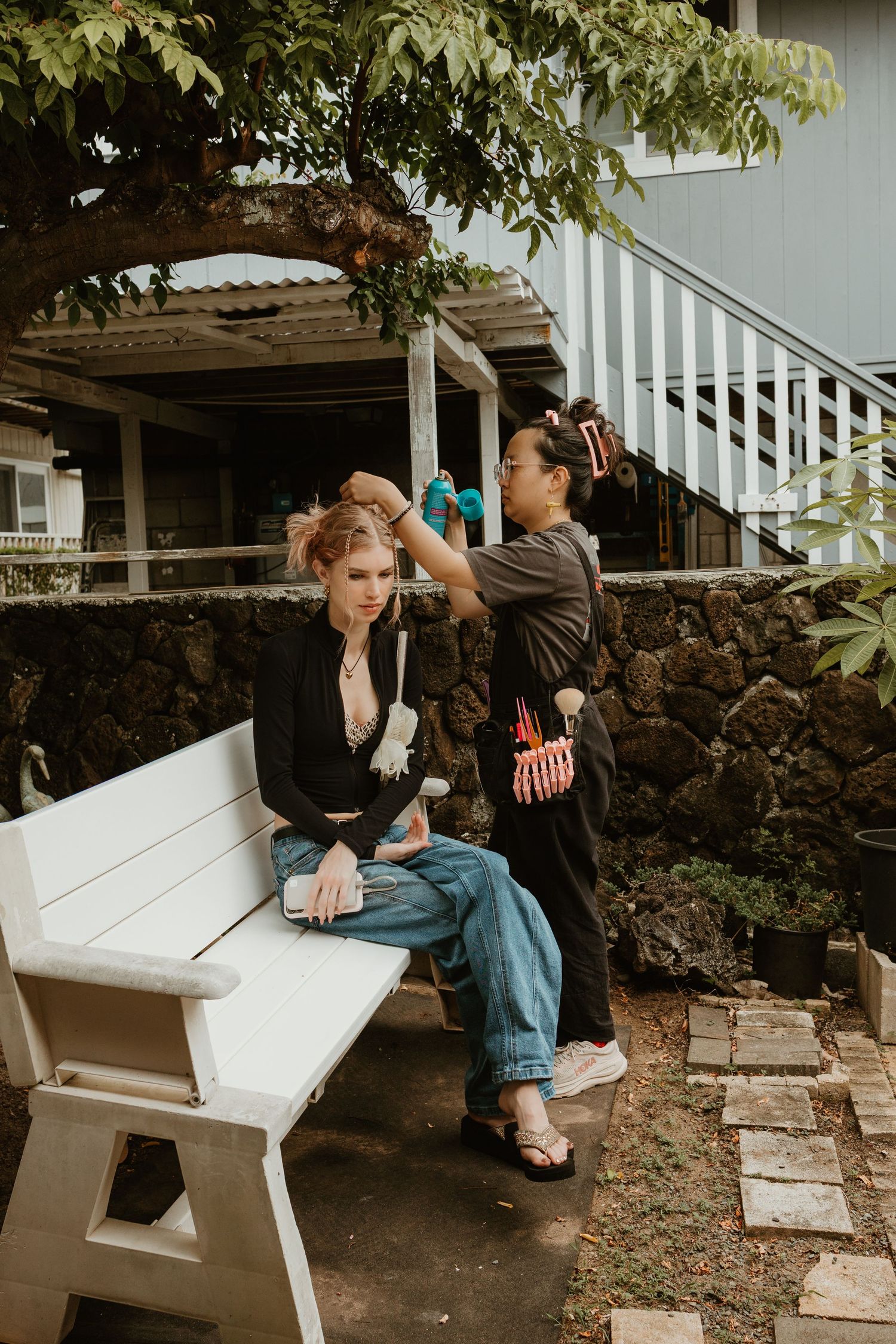 Two women outdoors, one sitting on a white bench while the other styles her hair near a stone wall.