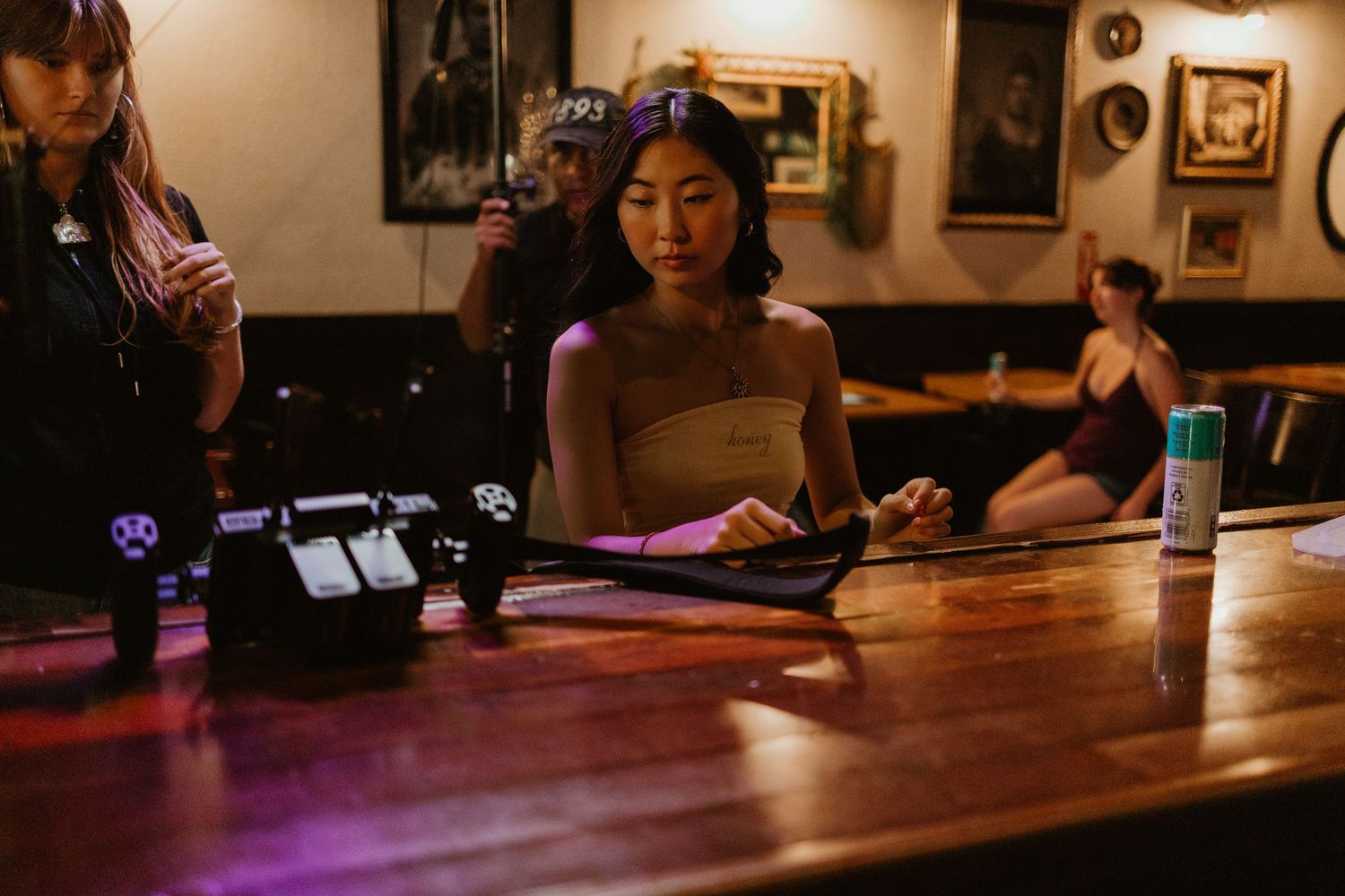 Young woman in strapless top writes at a wooden bar counter with guitar pedals nearby.