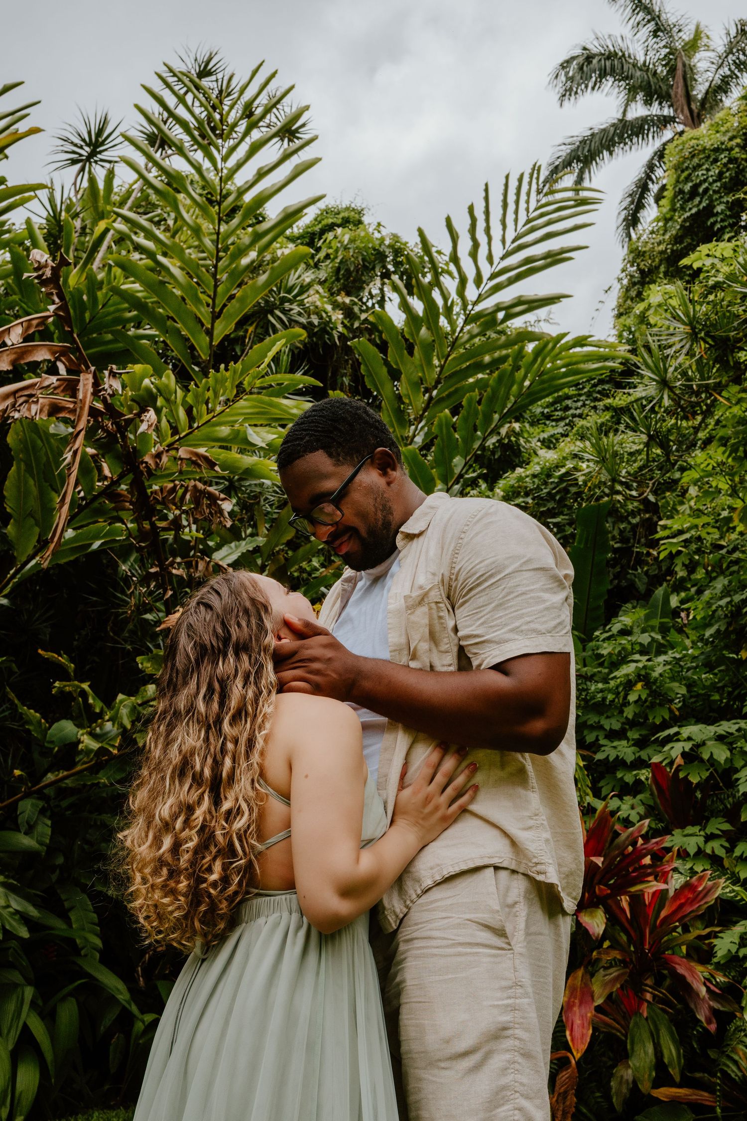 A couple shares a tender moment surrounded by lush tropical greenery, with the man gently holding the woman's face.