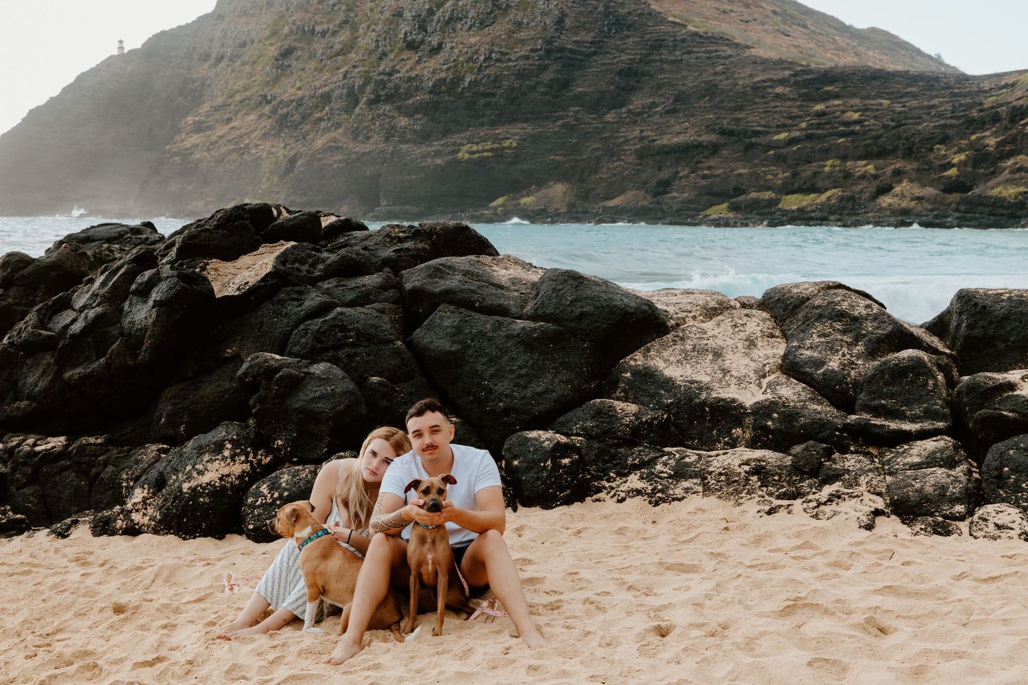 Couple sitting on sandy beach with rocky cliffs and ocean waves in Hawaii.