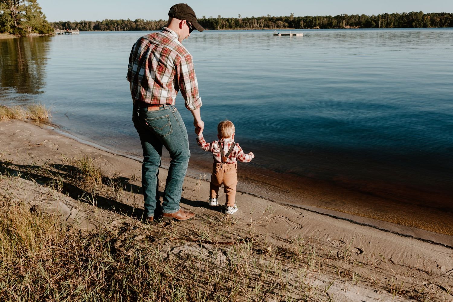 Father holds toddler's hand while walking along a sandy lakeshore surrounded by pine trees in warm autumn light.