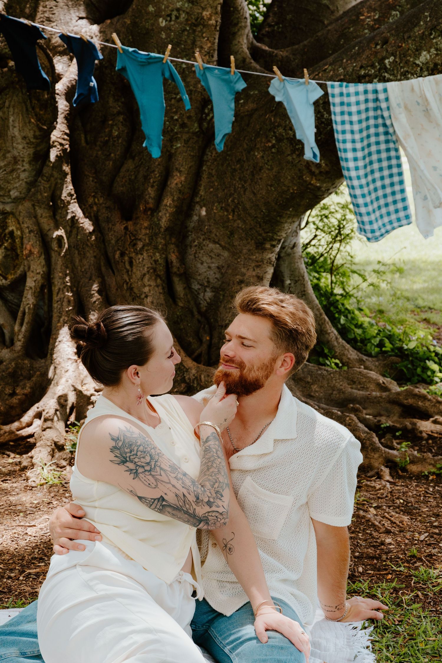 Couple sitting under large tree with blue bunting, woman touching mans beard affectionately.