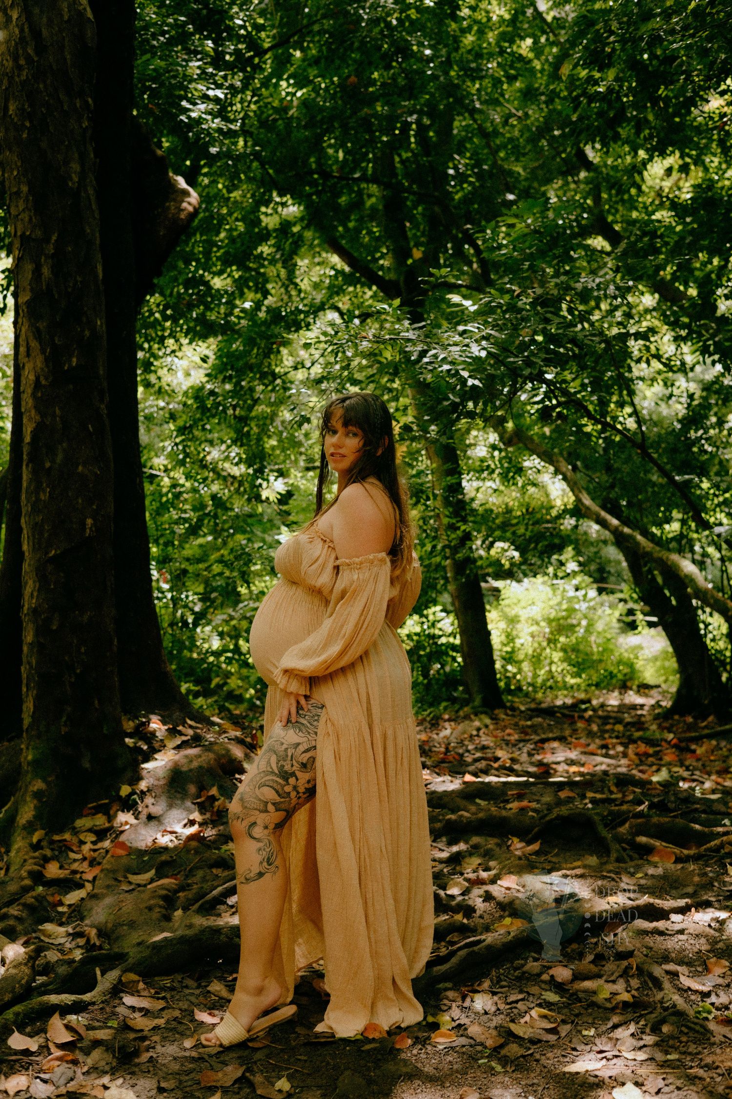 Pregnant woman in sheer gold maternity gown standing among lush green trees.