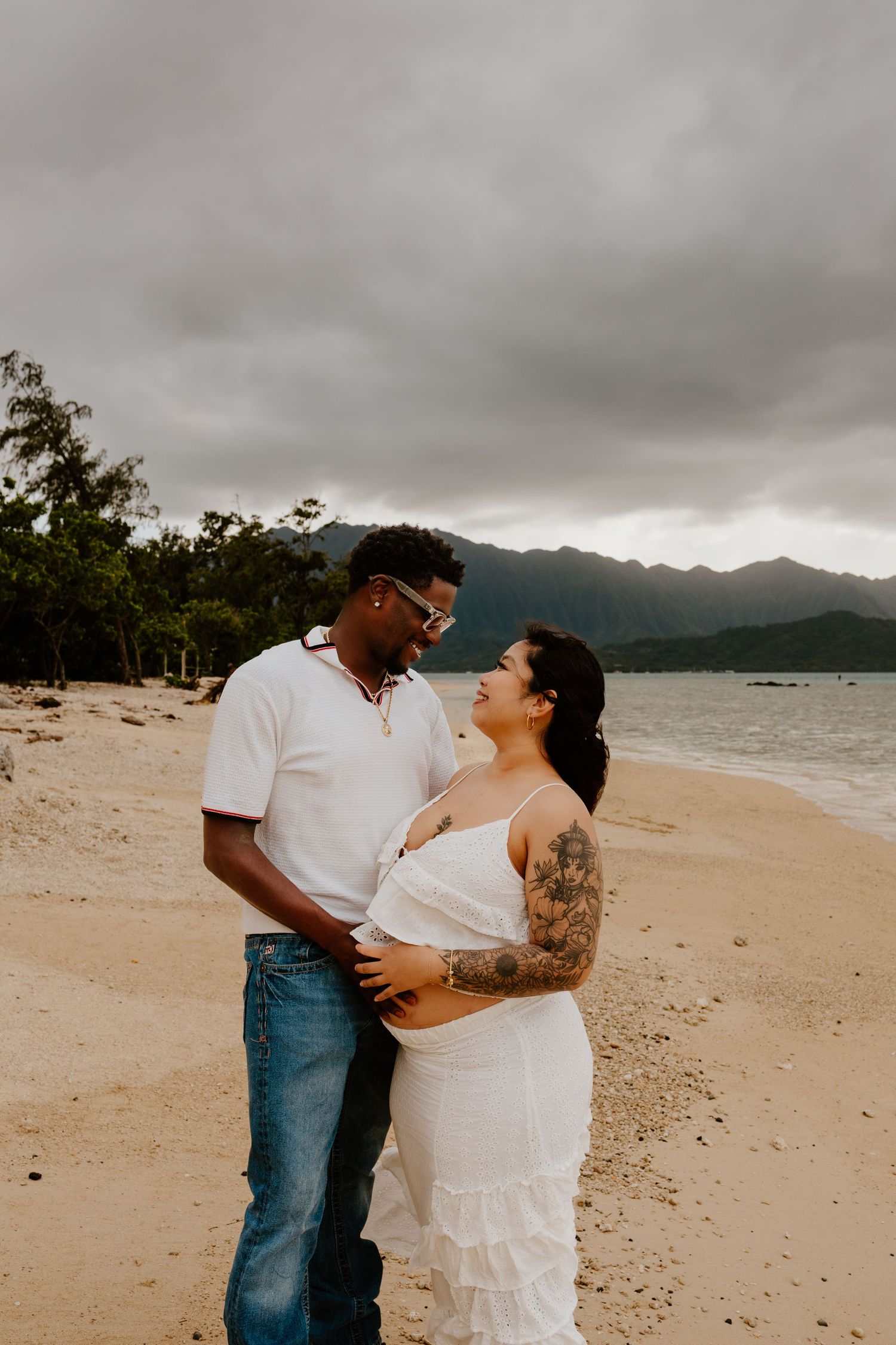 Interracial couple smiling together on sandy beach during maternity photoshoot.