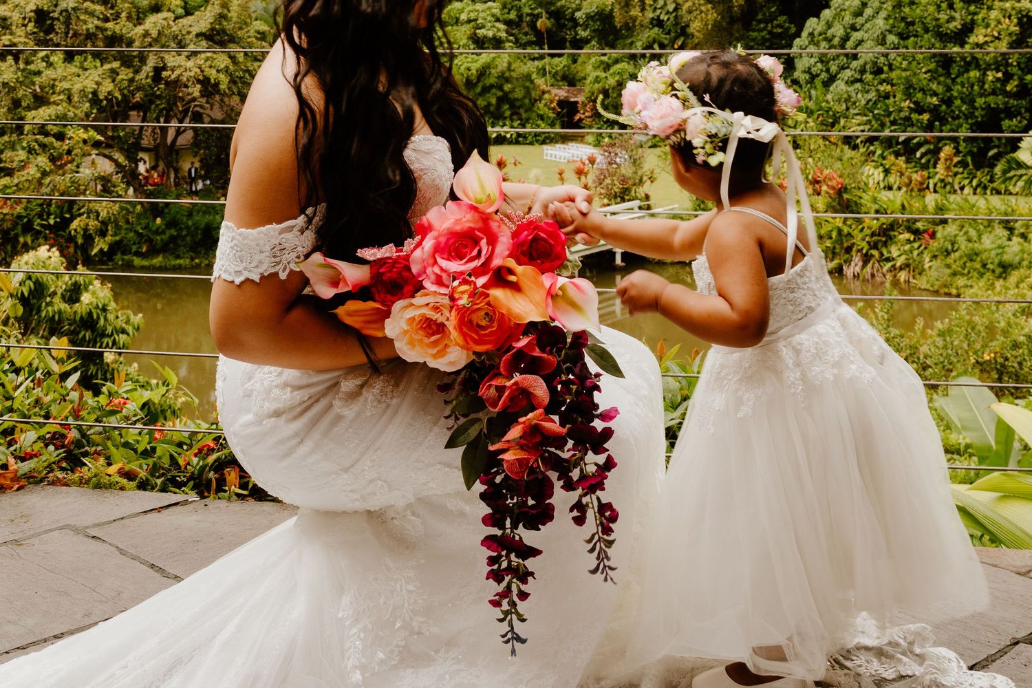 Bride in white off-shoulder gown holds vibrant floral bouquet as flower girl in white dress reaches up outdoors.