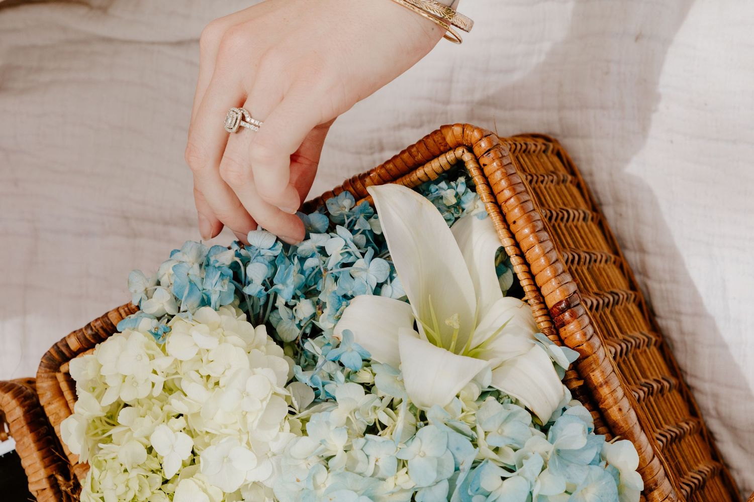 A hand with a diamond ring reaches into a wicker basket filled with blue hydrangeas and white lilies.