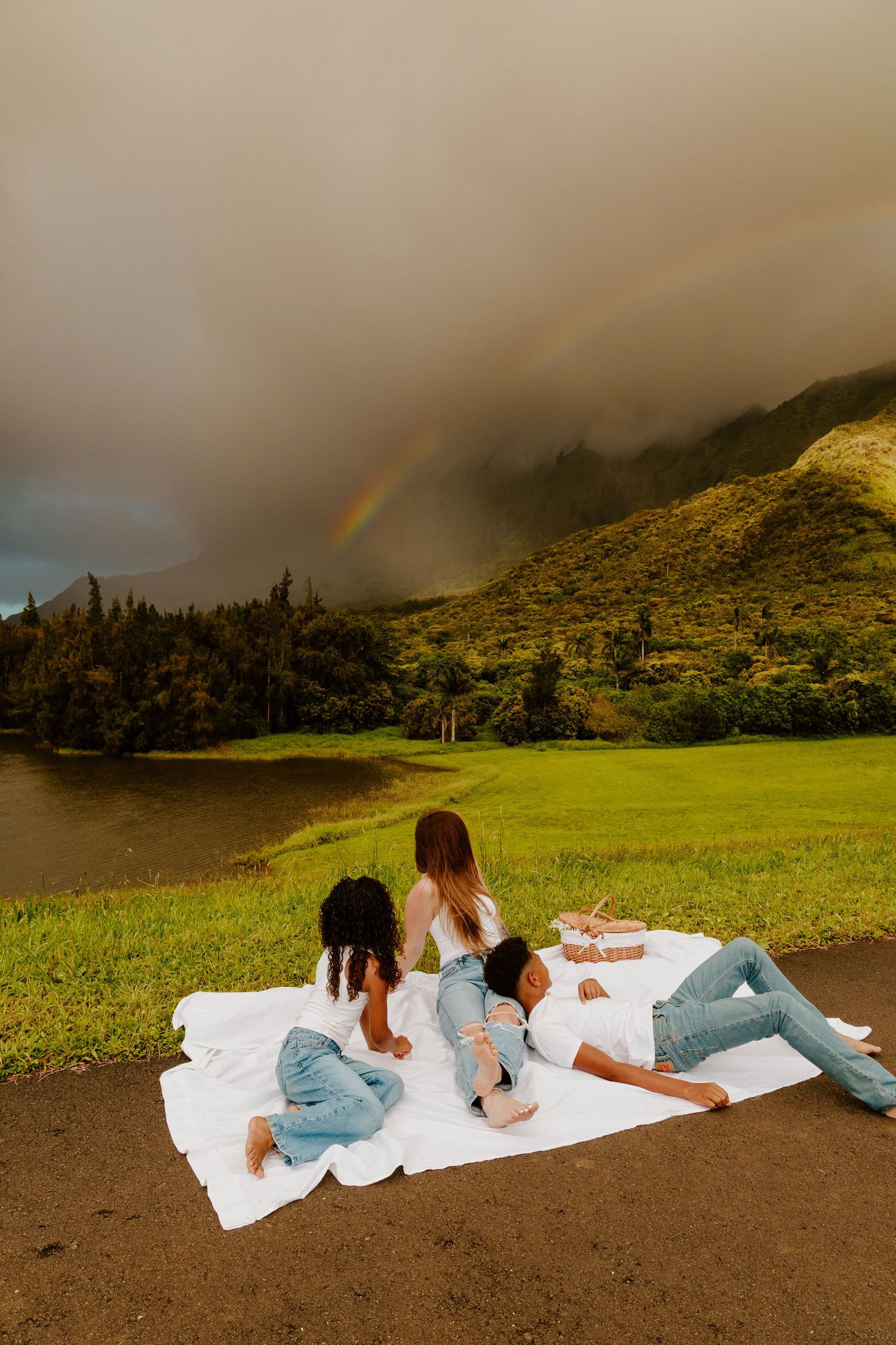Family sitting on a white blanket watching a vibrant rainbow over a scenic mountain lake.